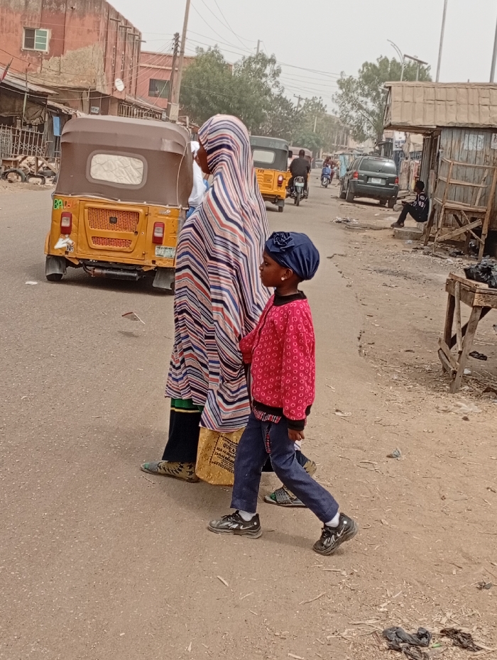 The image displays a street scene in Gawuna, Nigeria, under a hazy, light gray sky. In the foreground, a woman and a young girl walk towards the right on an unpaved, light brown road. The woman, viewed from the back-left, wears a full-length garment featuring horizontal stripes in blue, white, and reddish-brown over dark pants and patterned flat shoes. She carries a light-colored bag. The girl, positioned slightly ahead and to her right, wears a navy blue head covering with a rosette embellishment, a red long-sleeved top with white polka dots, dark blue jeans, and black Nike sneakers.
To the left, a yellow three-wheeled auto-rickshaw with a brown canopy is partially visible, facing away from the viewer. Further down the street, another yellow auto-rickshaw, a black sedan, and a person on a motorcycle are present. On the right side of the road, a makeshift wooden stall with a corrugated metal roof is visible, with a male individual seated on a concrete platform beside it. In the background, aged, multi-story buildings with reddish-brown facades and visible windows line the left side. Utility poles and overhead wires are present throughout the midground and background, and sparse trees are visible in the distance. Debris, including dark plastic or fabric, is present on the road surface in the foreground.