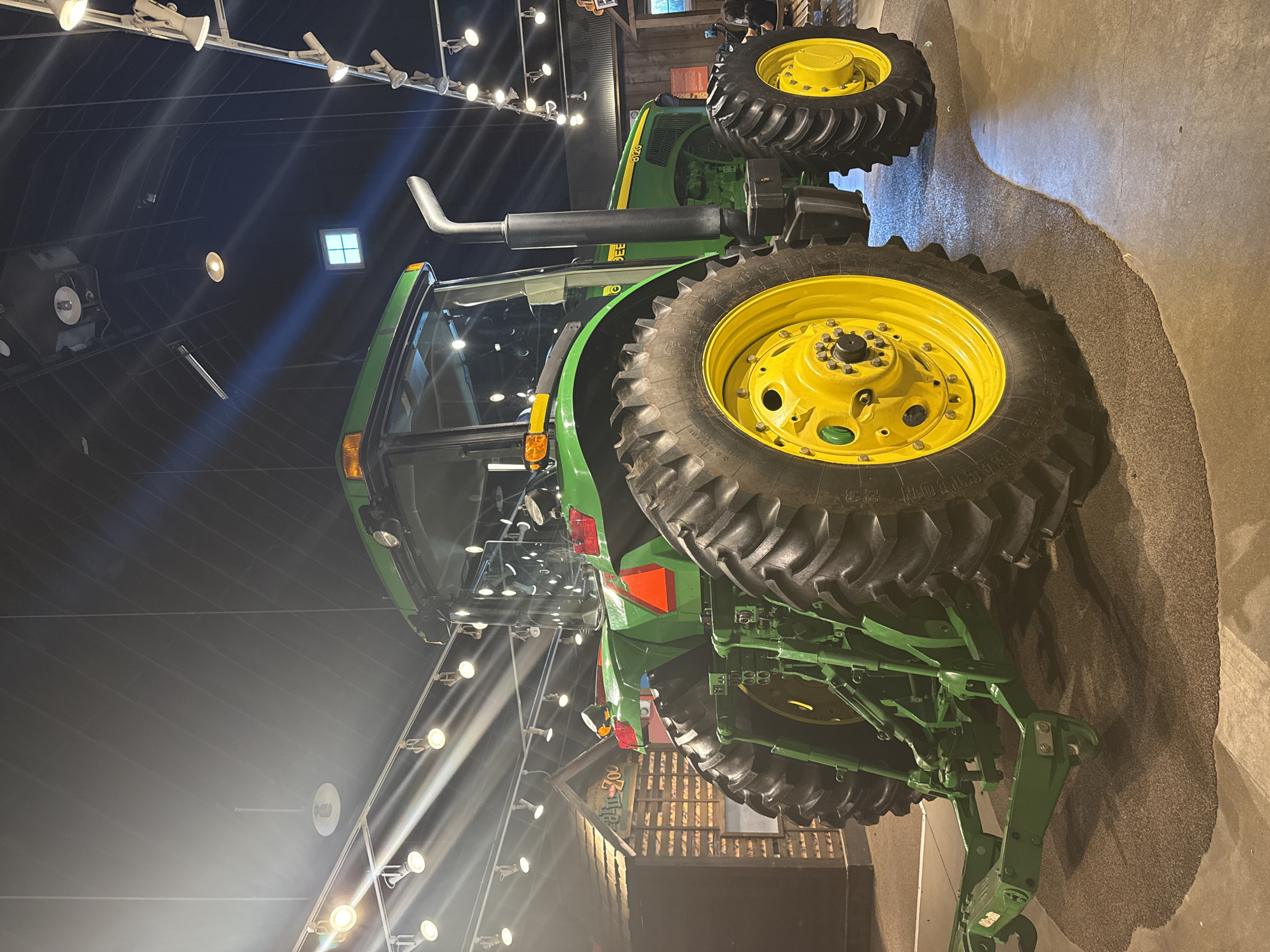 The image shows a green and yellow John Deere tractor. The tractor is located in an indoor setting, likely a museum or exhibit hall, given the presence of track lighting on the ceiling and a smoothed concrete floor. There is no evidence to confirm the location is Chicago, Illinois. The focus is on the tractor's features, including the large tires, exhaust pipe, and rear hitch assembly. The environment appears to be dimly lit, with the tractor illuminated by artificial lighting. No people are visible in the image.