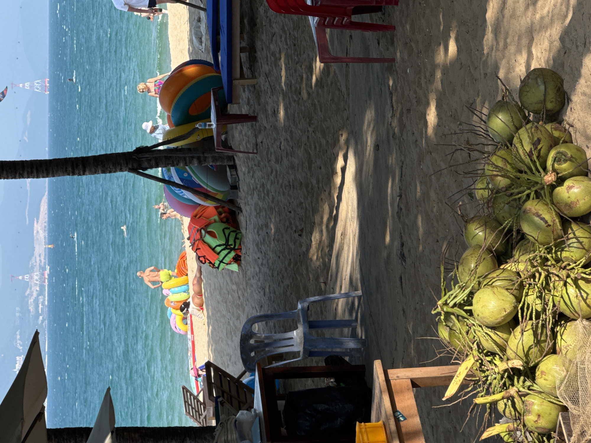 The image depicts a coastal scene, likely a beach, with various elements. A beach is visible, with numerous people engaging in water activities. The water is blue, and there are some structures or objects in the distance that seem to be related to water sports or recreation.
On the right side of the frame, there's a large bunch of green coconuts. Various beach-related items are present, including inflatable rings and beach toys. In the foreground, there's furniture like chairs and tables, suggesting a beachside cafe or resting area. The location is unknown.