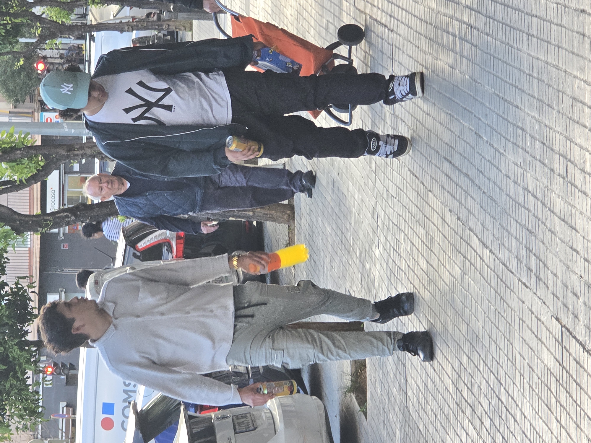The image shows an urban scene in Cornellà de Llobregat, Spain. There are two young men and one older man. The first young man, on the right, wears a white long-sleeved shirt and light-colored pants. He holds a can and a yellow and orange object in his hands. The second young man wears a light green NY Yankees cap, a white t-shirt with the NY Yankees logo, a black jacket, and black pants, also holding a can in his hand. An older man stands between them, dressed in a blue jacket and dark pants. There is a stroller near the young man in the cap. The scene takes place on a paved street with trees in the background, and a car and a commercial van are also visible.