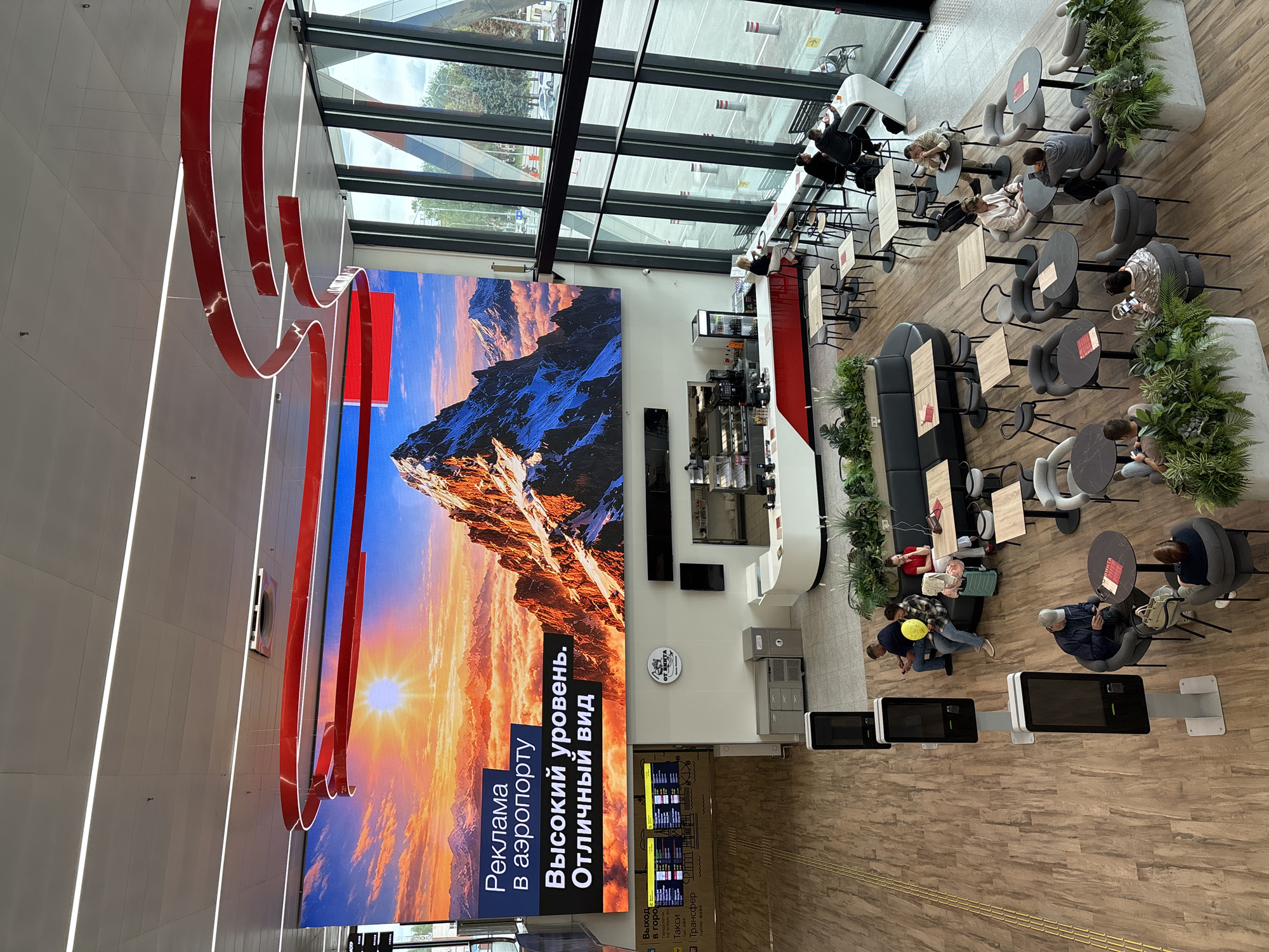 The image depicts the interior of an airport with a large digital screen displaying an advertisement for "Advertising at the airport: High level. Excellent view." The screen shows a mountain landscape with a sunset. Below the screen, a cafe area is visible with seating and tables. Several people are seated at tables in the cafe, some interacting with each other and others looking at their phones.
To the left of the cafe, a departure board is displayed, showing flight information. Further to the left, there are self-service kiosks. The floor is a light-colored wood laminate, and the ceiling features modern lighting fixtures and red decorative elements. Large windows on the right side of the image offer a view of the exterior, including trees and part of a parking lot.
The location is identified as the city of Старое Мартьяново, Russia, which is a small village in the Republic of Bashkortostan, Russia, and it is highly unlikely that this airport is located in such a small settlement. Therefore, the stated location is likely incorrect.