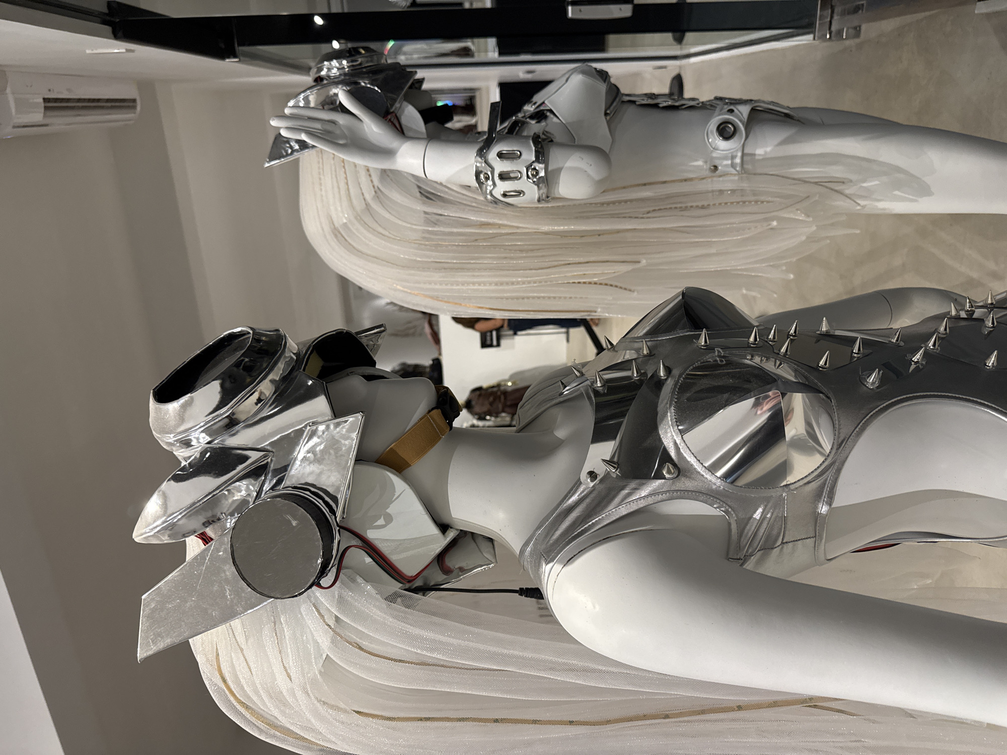 Two mannequins, displayed upright in what appears to be a gallery, showroom, or retail space, wear elaborate, futuristic, or cyberpunk-inspired outfits. The scene is likely indoors, possibly in Baan Lipa Noi, Thailand, given the context.
The mannequin in the foreground, positioned to the left, features a gleaming silver-chrome headpiece with angular, wing-like elements and a circular disc on the side. Red and black wires are visible, connecting to the headpiece. It wears a silver bra-top adorned with numerous sharp spikes. From the waist down, the mannequin is draped in a flowing, voluminous skirt made of translucent, white, sheer fabric, creating a layered, ethereal effect. A subtle golden band is visible around its neck.
The second mannequin, standing slightly behind and to the right, wears a similar silver-chrome headpiece and a prominent silver cuff or brace on its left wrist. This mannequin also features a long, flowing skirt of white, sheer, layered fabric, mirroring the style of the first. Its torso appears to have a silver-colored top or armor piece.
The background shows plain light-colored walls, with an air conditioning unit mounted high on the left wall. Reflections in the shiny surfaces of the costumes and other elements suggest a well-lit indoor environment, possibly revealing faint glimpses of other individuals or displays in the room. No discernible text is visible. The overall aesthetic is striking and modern, focusing on metallic and translucent textures.