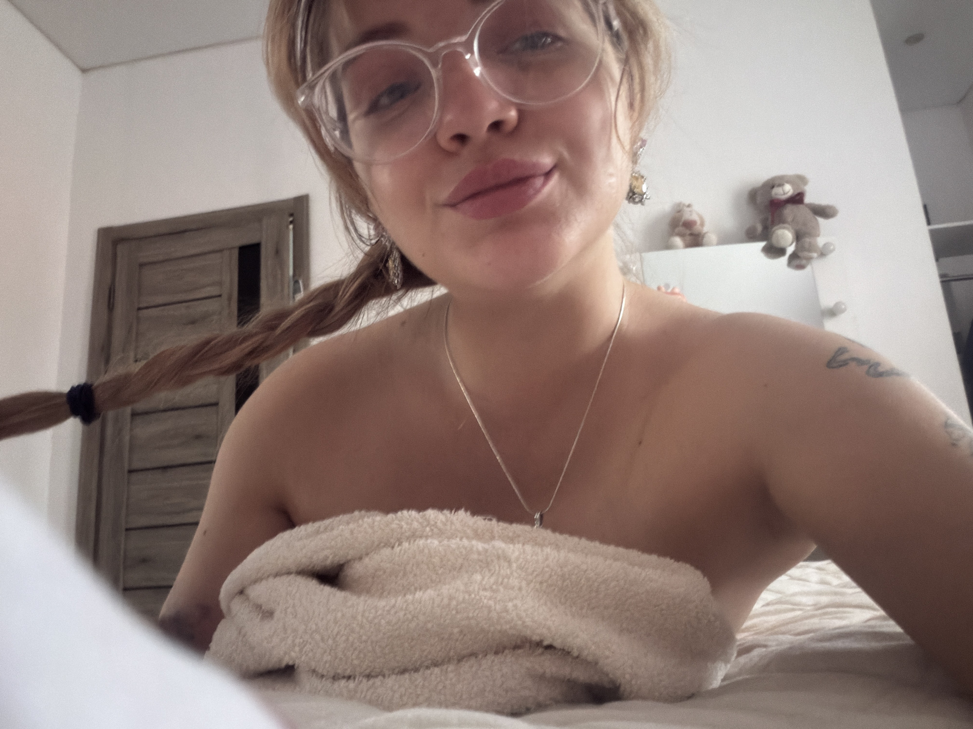 A blonde woman with glasses, hair in a braid, and a necklace is partially covered with a white towel. She is lying on a bed in a room with a wooden door and stuffed animals on top of a white dresser in the background. The location may be in Большая Шильна, Russia.