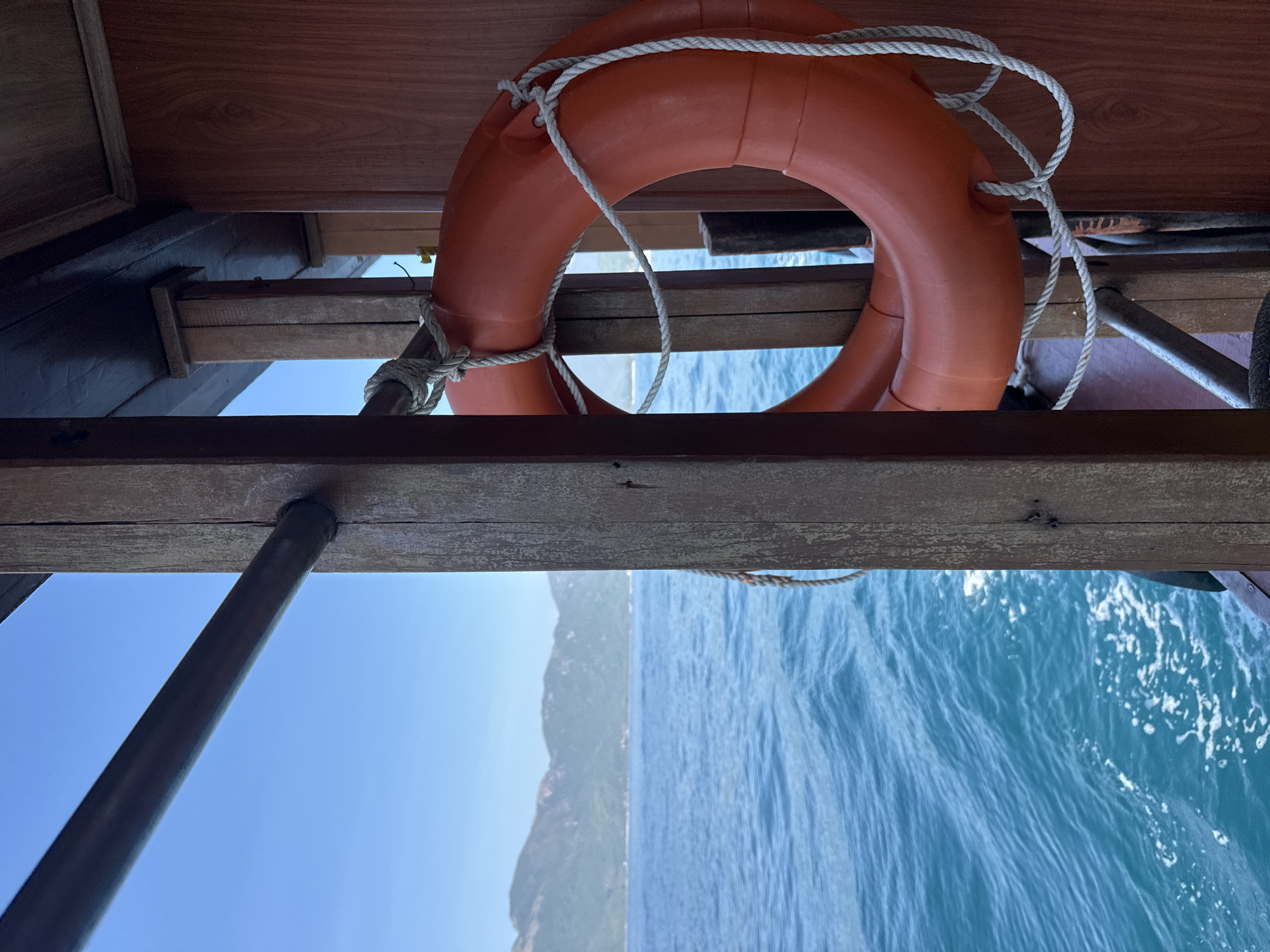The image is taken from a boat in the waters off the coast of Nha Trang, Vietnam. In the foreground, a wooden structure of the boat is visible, with a life preserver hanging from a rope on a wooden post. The life preserver is bright orange and made of a buoyant material. To the left of the boat's structure, the turquoise waters of the sea are visible, with gentle waves and white foam. In the distance, a range of green, mountainous islands can be seen against a clear blue sky. The scene captures a moment of travel and the natural beauty of the coastal landscape.