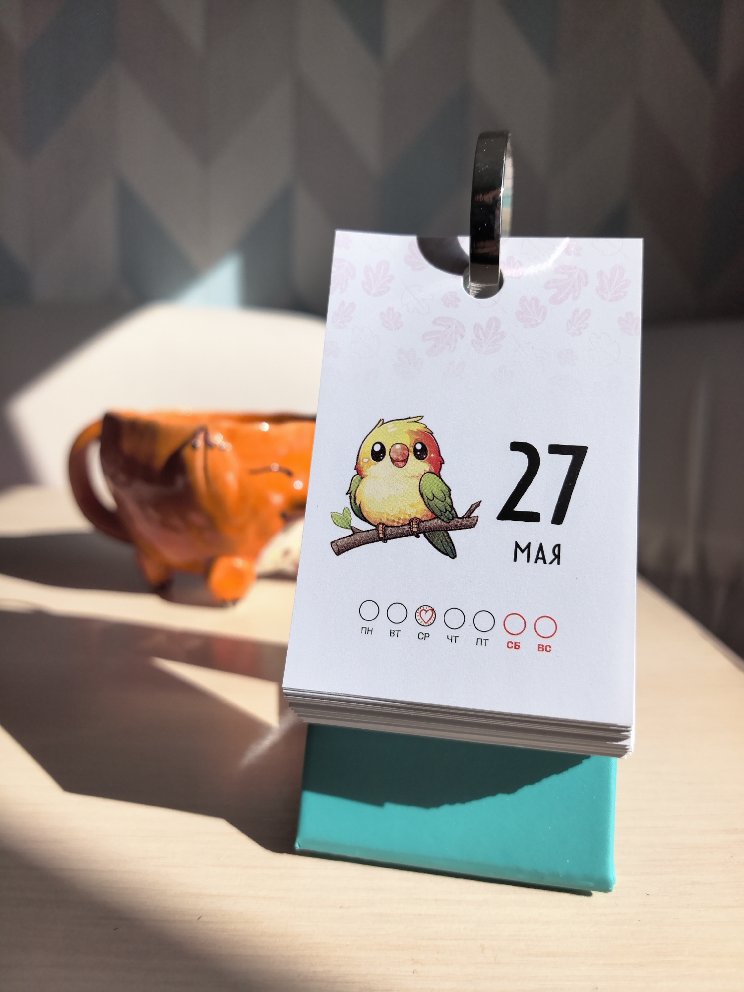 A close-up photograph shows a desk calendar displaying the date "27 МАЯ", indicating May 27th. Above the date is a cartoon illustration of a colorful bird perched on a branch. Below the date are symbols for the days of the week, with the weekend days circled in red. Behind the calendar, out of focus, is an orange fox-shaped mug. The calendar rests on a turquoise base. The lighting suggests an indoor setting with sunlight. No people are visible in the image. The location is specified as Фролы, Russia.