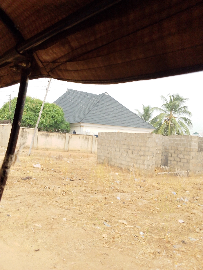 This image depicts an outdoor scene in Ajaokuta, Nigeria, observed from under a brown fabric canopy, likely part of a vehicle's roof, which occupies the top third of the frame. A dark metal support pole for the canopy is visible on the left. The immediate foreground shows dry, sandy-brown soil sparsely covered with dead yellowish vegetation and scattered white and dark litter.
In the mid-ground, a light-colored concrete block wall, approximately 1.5 meters high, stretches horizontally from left to center. Behind this wall, on the left, a tall wooden utility pole stands, and a dense, full-canopied green tree, possibly a mango tree, is visible. To the right of the scene, an unfinished structure made of gray concrete blocks forms a partial wall, about waist-height, with an apparent opening for a door or window.
The background is dominated by a modern, single-story residential building with light-colored walls and a complex, multi-gabled roof covered in dark gray or black tiles. The building appears to be enclosed by a fence topped with barbed or razor wire. To the right of this building, a tall palm tree with green fronds and visible coconuts stands against an overcast, light gray sky. No individuals are present in the scene.