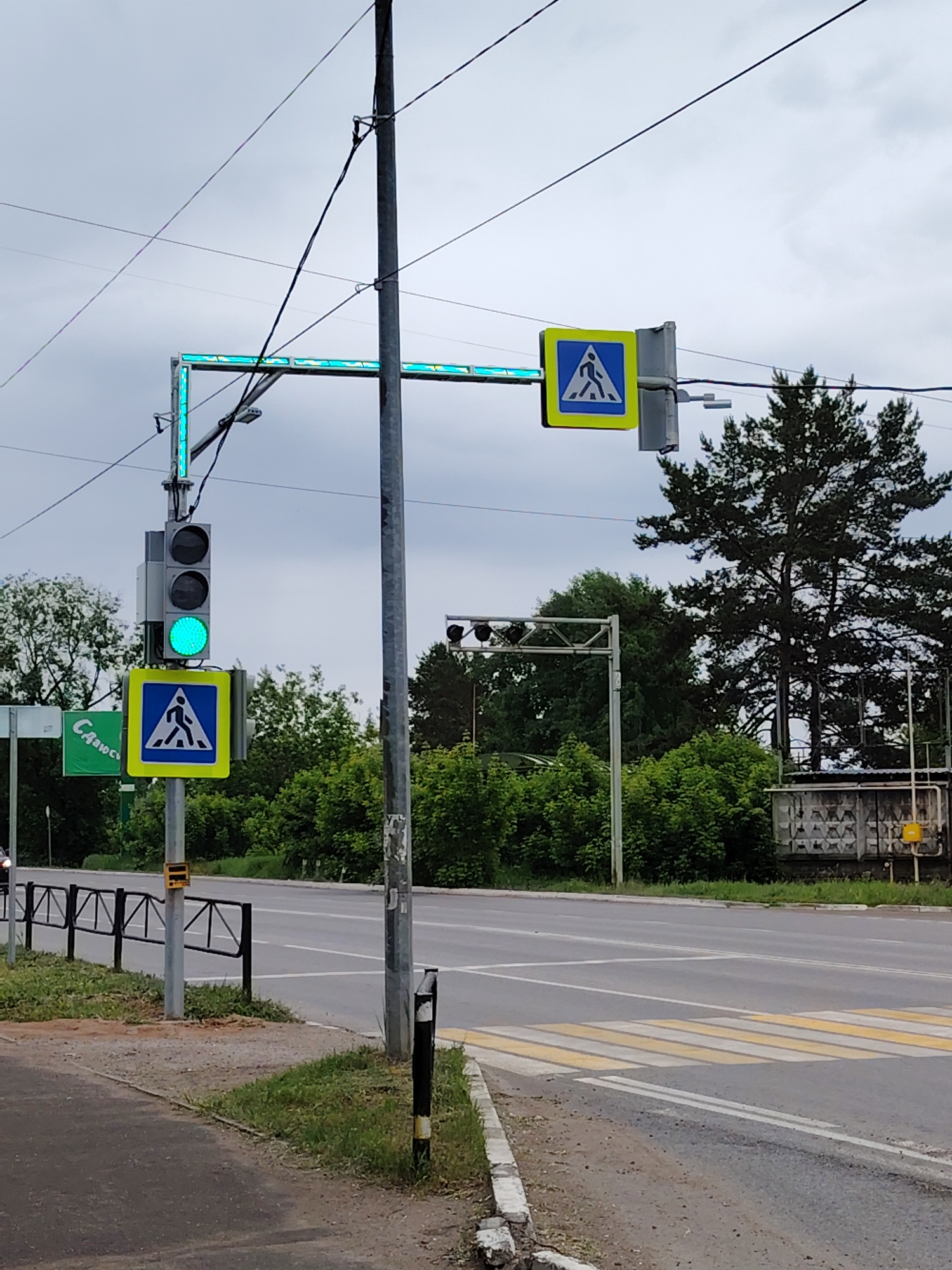 This daytime image, taken in Chaikovsky, Russia, depicts a pedestrian crossing under an overcast sky. In the foreground, a paved sidewalk abuts a low black metal fence that runs parallel to a road. The road is clearly marked with prominent yellow and white striped zebra crossing lines.
Central to the scene is a utility pole supporting a vertical traffic light, which displays a green signal on its lowest lamp, indicating permission to proceed. Below this traffic light, a standard pedestrian crossing sign – a blue triangle with a white pedestrian icon on a yellow square – is clearly visible. An identical pedestrian crossing sign is mounted overhead on a gantry structure. This gantry is equipped with integrated green LED lighting, illuminating brightly and extending horizontally across the road, visually emphasizing the crossing area.
Numerous utility wires crisscross the upper part of the frame, connected to the primary pole. In the background, lush green trees and dense foliage line the road. Further back on the right, there's a grey building, possibly industrial or a utility structure, with visible pipes and a yellow circular fixture. A partially visible green sign on the far left displays Cyrillic text that appears to start with "Сдаю...", likely part of a "For Rent" advertisement. The image captures a functional urban intersection with active traffic management for both vehicles and pedestrians.