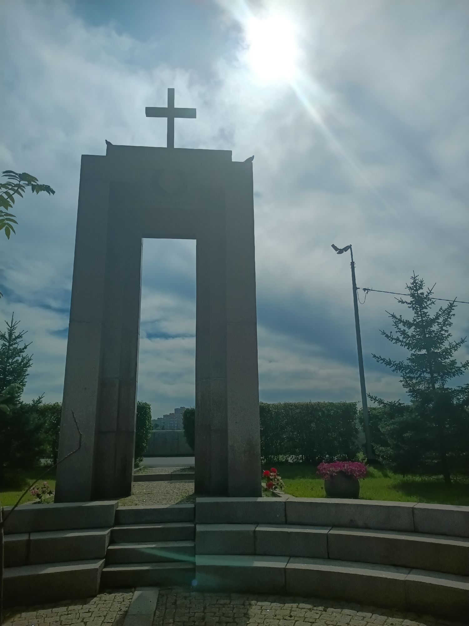 The image depicts the Memorial to the Victims of Political Repression in Irkutsk, Russia. A large, gray stone archway stands prominently, topped with a cross. The sun shines brightly through a cloudy sky, creating lens flare. Two birds are perched on top of the arch. In front of the archway, tiered stone steps lead upwards. Beyond the arch, a neatly trimmed hedge forms a backdrop, with evergreen trees and a lamppost visible. Two potted plants with red and pink flowers are placed on the grass to the right of the steps. The ground is paved with cobblestones. The overall scene is a somber and respectful memorial site under a partly cloudy sky.