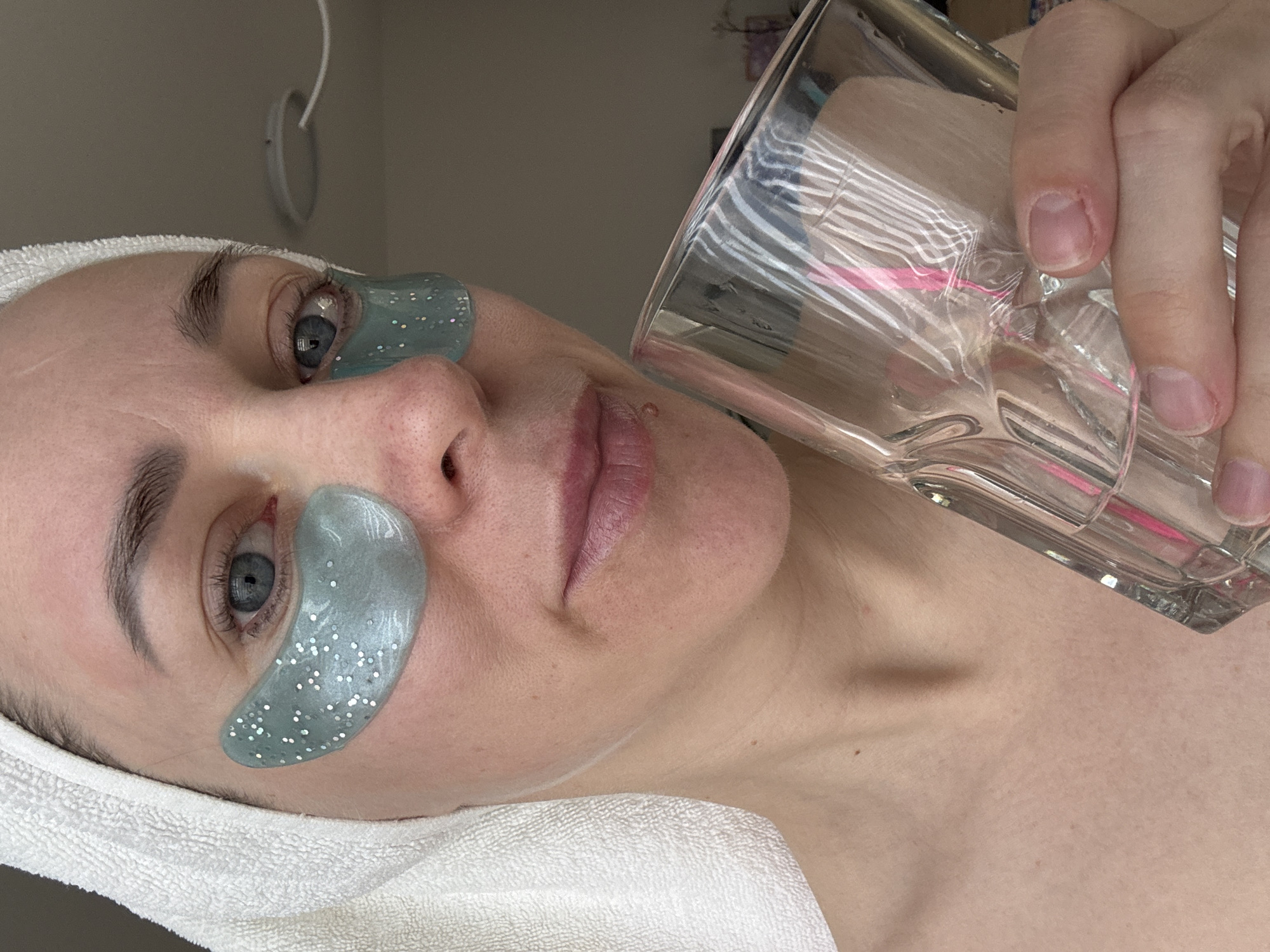 A fair-skinned person is lying down, facing upwards. They have a white towel wrapped around their head. Underneath each eye is a glittery, teal-colored eye mask. They are holding a clear glass in their right hand. The glass appears to be empty. The background is a light, neutral color. The location in the city of Chaikovsky, Russia, cannot be determined from the image.