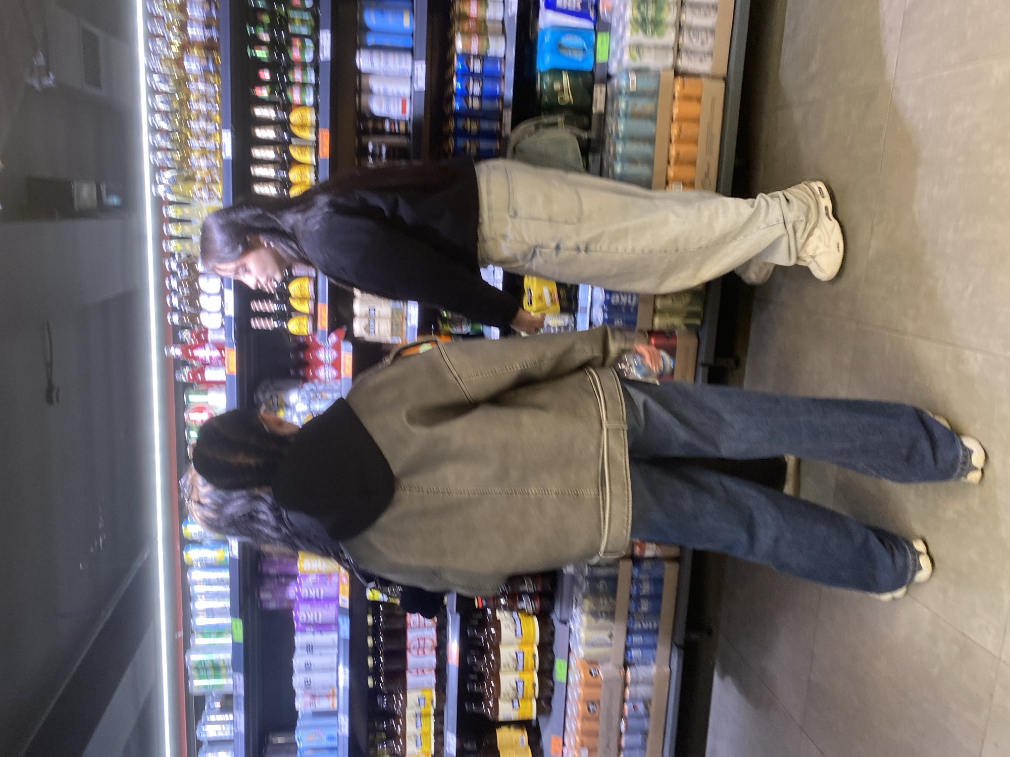 The image captures an indoor scene within a supermarket or convenience store aisle in Poltava, Ukraine, viewed from a slightly elevated and rotated perspective. Two young women are the main subjects, engaged in shopping for beverages.
The person positioned further back on the left has long dark hair and wears a dark long-sleeved top, light-colored wide-leg pants, and white sneakers. She is leaning towards the refrigerated shelves, appearing to examine items.
The second person, closer to the foreground and on the right, wears a dark hoodie under an olive or khaki-colored jacket with lighter trim, dark blue wide-leg jeans, and white or cream-colored chunky sneakers. She is also facing the shelves, seemingly browsing the products, and her hand appears to be holding a small bottle or can.
The background consists of tall, brightly lit refrigerated shelves densely stocked with various bottled and canned beverages. Prominently visible on numerous yellow-labeled bottles is the Cyrillic text "ОБОЛОНЬ," indicating the Ukrainian brand Obolon. Other products feature blue, red, and purple labels; some purple-labeled cans might show "1KG" or similar text, though it is blurry. The floor is a light gray tiled surface. The bright lighting suggests a typical retail environment during operating hours.