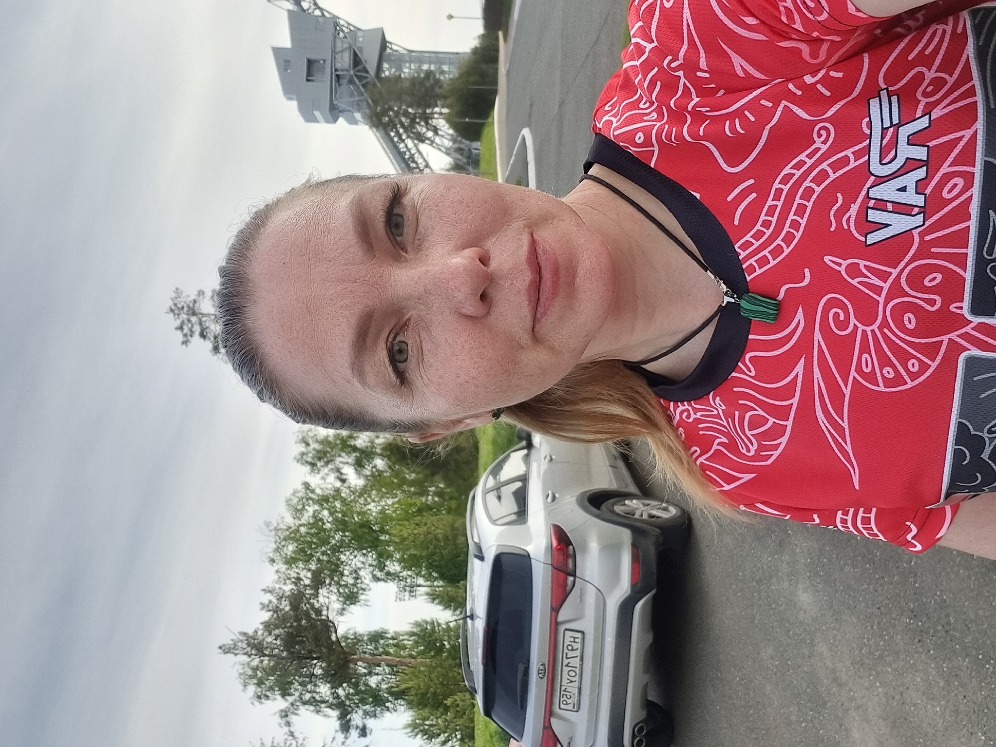 A woman with fair skin, freckles, and light eyes is facing the camera. She is wearing a red jersey with white graphic patterns and black trim. A black cord necklace with a green pendant is visible around her neck. Her blonde hair is partially pulled back.
Behind her is a silver-colored SUV. The license plate reads "Р237ОГ159" in Cyrillic script, indicating it is registered in Russia.
In the background, there are trees and an industrial structure resembling a tower or crane with scaffolding. The photo appears to have been taken in an outdoor location near a roadway or parking area. Based on the license plate, the location is in the city of Прикамский, Russia.
