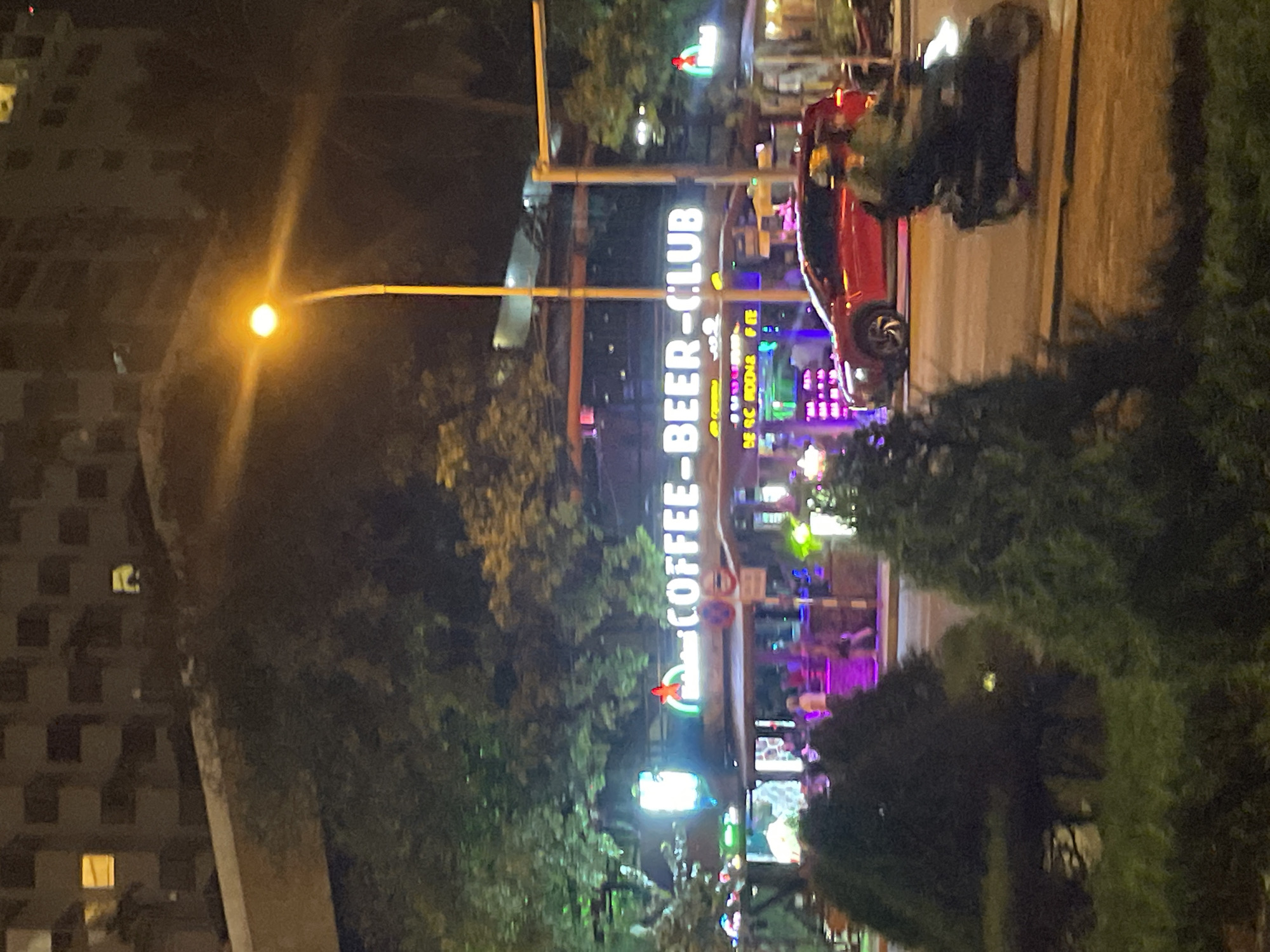 The image depicts an outdoor scene at night in Nha Trang Ward, Vietnam. Dominating the view is a vertically oriented neon sign reading "COFFEE-BEER-CLUB". The sign illuminates a street area, where people are present, though specific actions and interactions are not discernable due to distance and image quality. A red car is parked nearby. Dense foliage is visible in the foreground and partially obscures the street. To the left, a high-rise building is partially visible, with a few lit windows. A street lamp is present, with its light creating a noticeable halo.