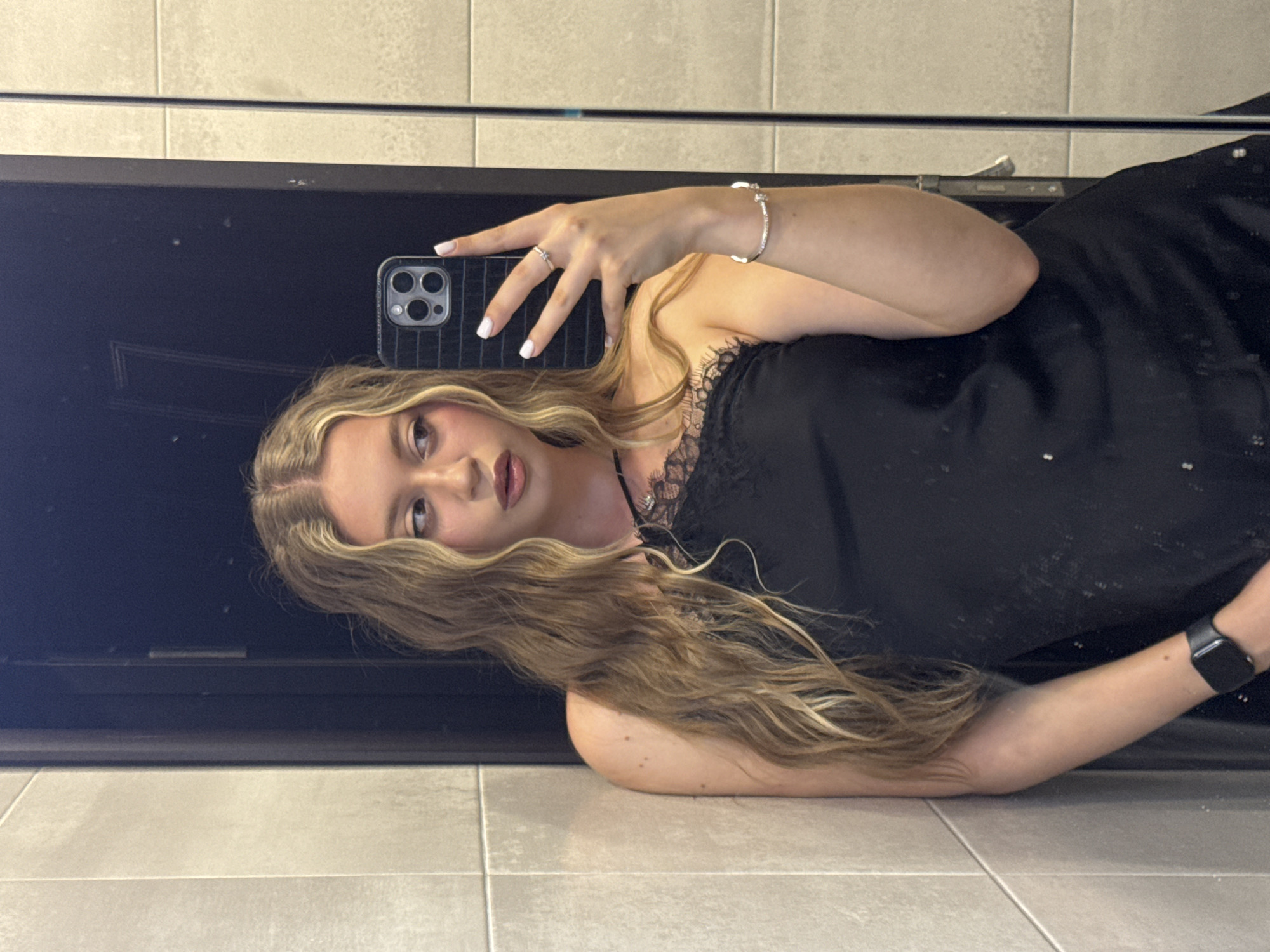 A young woman with long, wavy blonde hair is lying on a tiled floor and taking a mirror selfie. She is wearing a black dress with lace trim. On her left wrist, she is wearing a smartwatch, and on her left hand, she has a ring. Her right hand is holding a phone, which is angled to capture her reflection. The background consists of a tiled wall and a dark surface, likely a mirror or a piece of furniture. The lighting suggests it could be daytime, as there is no indication of artificial light sources. The woman's expression is neutral, and the overall scene is casual and personal. No visible text is present in the image.