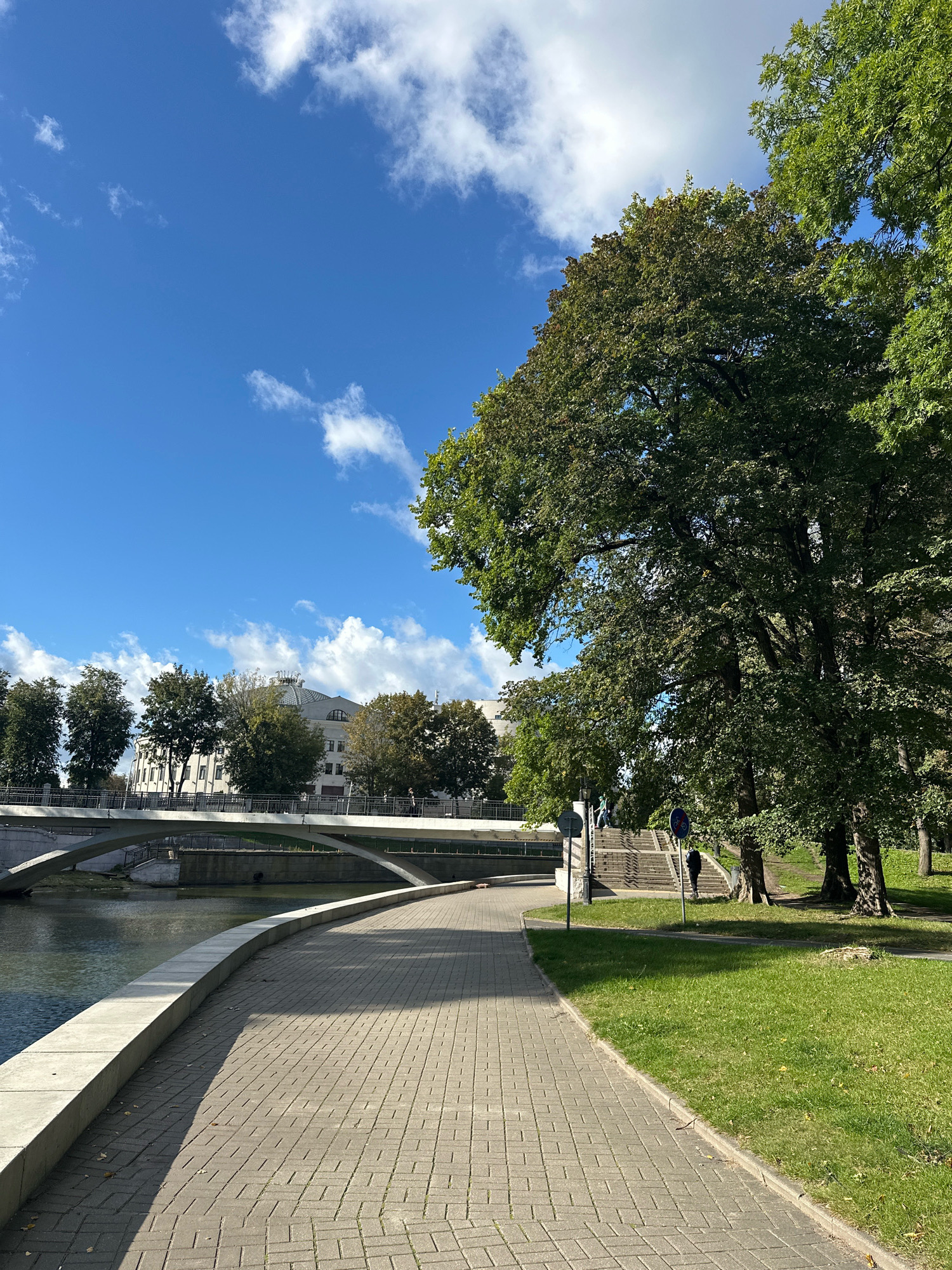 The image depicts a scenic view of the Svislach River embankment in Minsk, Belarus, on a clear day with scattered white clouds. A paved walkway with a stone curb lines the river. Across the river, a modern bridge with concrete arches spans the water. Beyond the bridge, buildings and trees are visible. To the right of the walkway, a well-maintained grassy area is shaded by large, leafy trees. In the mid-ground, a set of stairs leads up from the embankment, and a few individuals are seen walking or standing. The overall scene suggests a public park or recreational area.