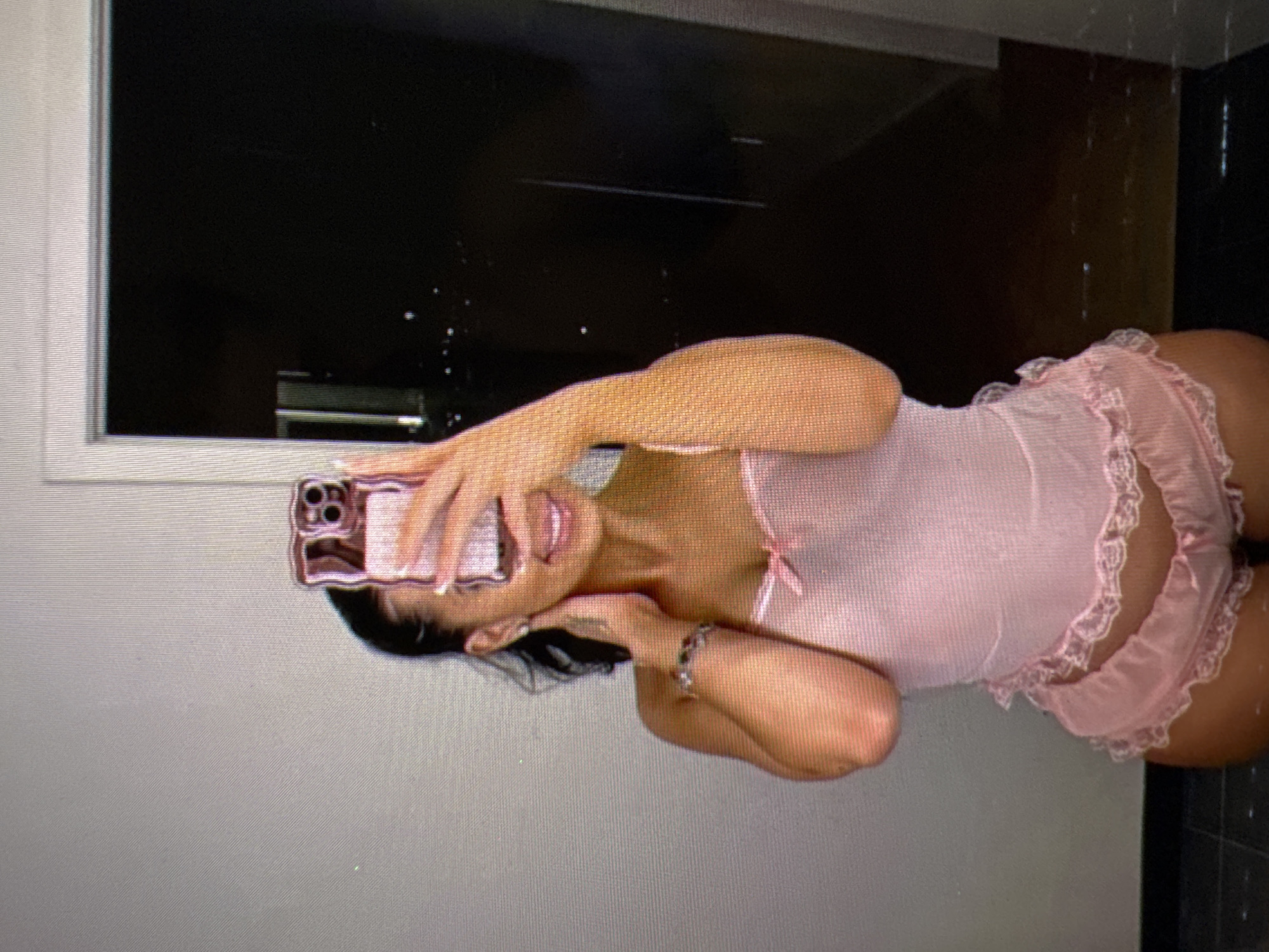 A woman is taking a mirror selfie in a room. She is wearing a pink lace camisole and matching shorts. Her hair is dark and pulled back. She is smiling and holding a pink phone in her right hand. The room appears to be a bedroom or dressing area, with a gray wall and a doorway in the background. There is a black floor with a tiled pattern. The image does not contain any information that allows for the identification of its location in Castelo Branco, Portugal.