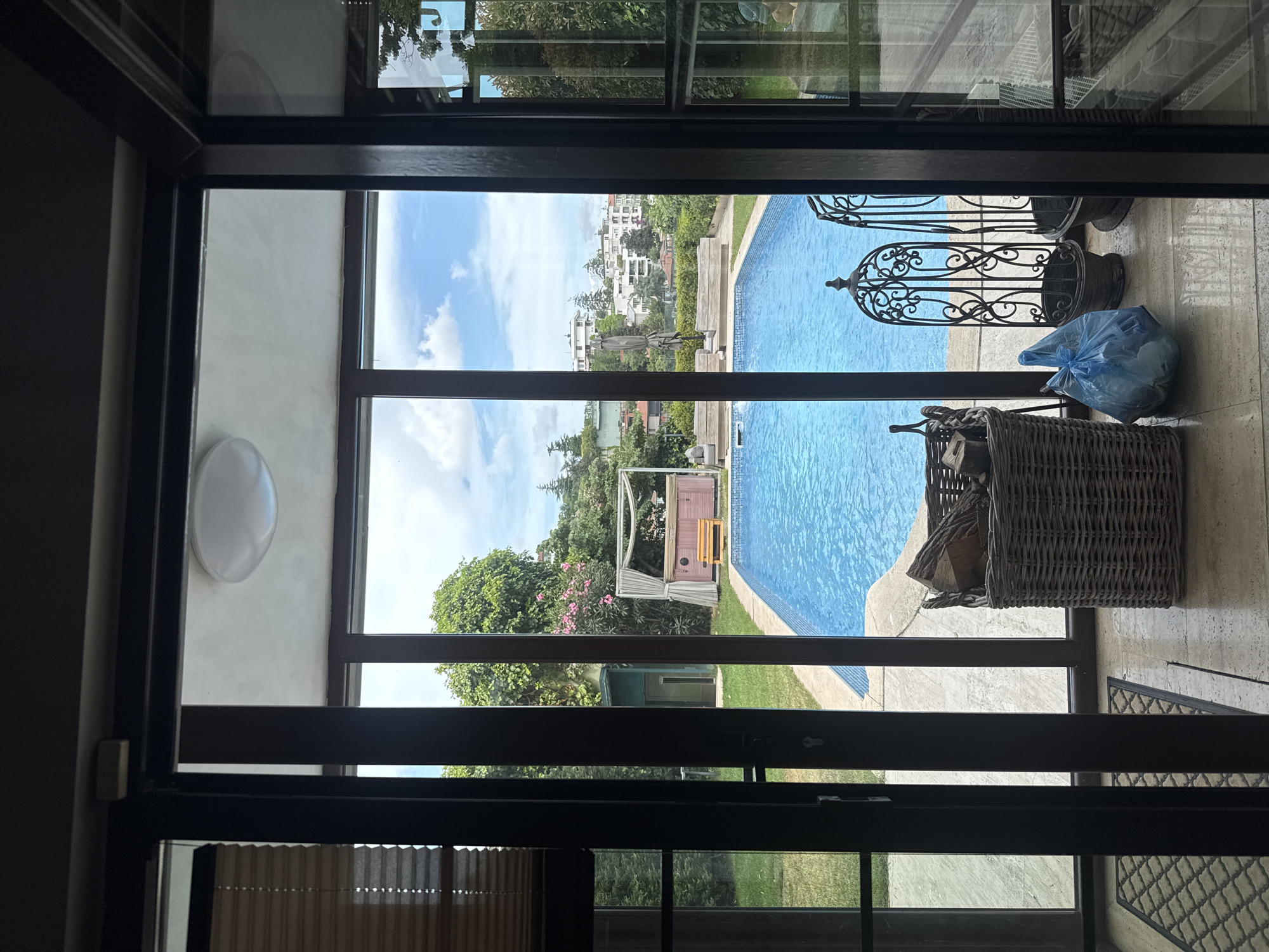 The image shows a view through an open doorway onto an outdoor scene. A bright blue swimming pool dominates the right side of the frame, with a tiled edge and a light-colored patio surrounding it. Beyond the pool, there is a lush green lawn and trees. In the distance, several white buildings are visible on a hillside, suggesting an urban or suburban setting.
On the patio, several objects are present. A large wicker basket filled with logs sits near the pool's edge. Further to the right, a blue plastic bag is tied shut, and next to it are two ornate black metal stands, possibly decorative planters or candle holders. A small pink gazebo-like structure with white curtains is visible on the lawn, adding a touch of whimsical design to the outdoor space. The sky is blue with scattered white clouds, indicating fair weather.
The perspective suggests the photo was taken from inside a building, looking out through a large window or door frame. The interior visible in the foreground is dimly lit and features dark-colored framing. The overall impression is of a pleasant, sunny day at a residential or resort-like location. The image does not contain any visible text. The location context provided, Beşiktaş, Turkey, aligns with the Mediterranean architectural style of the distant buildings and the lush greenery.