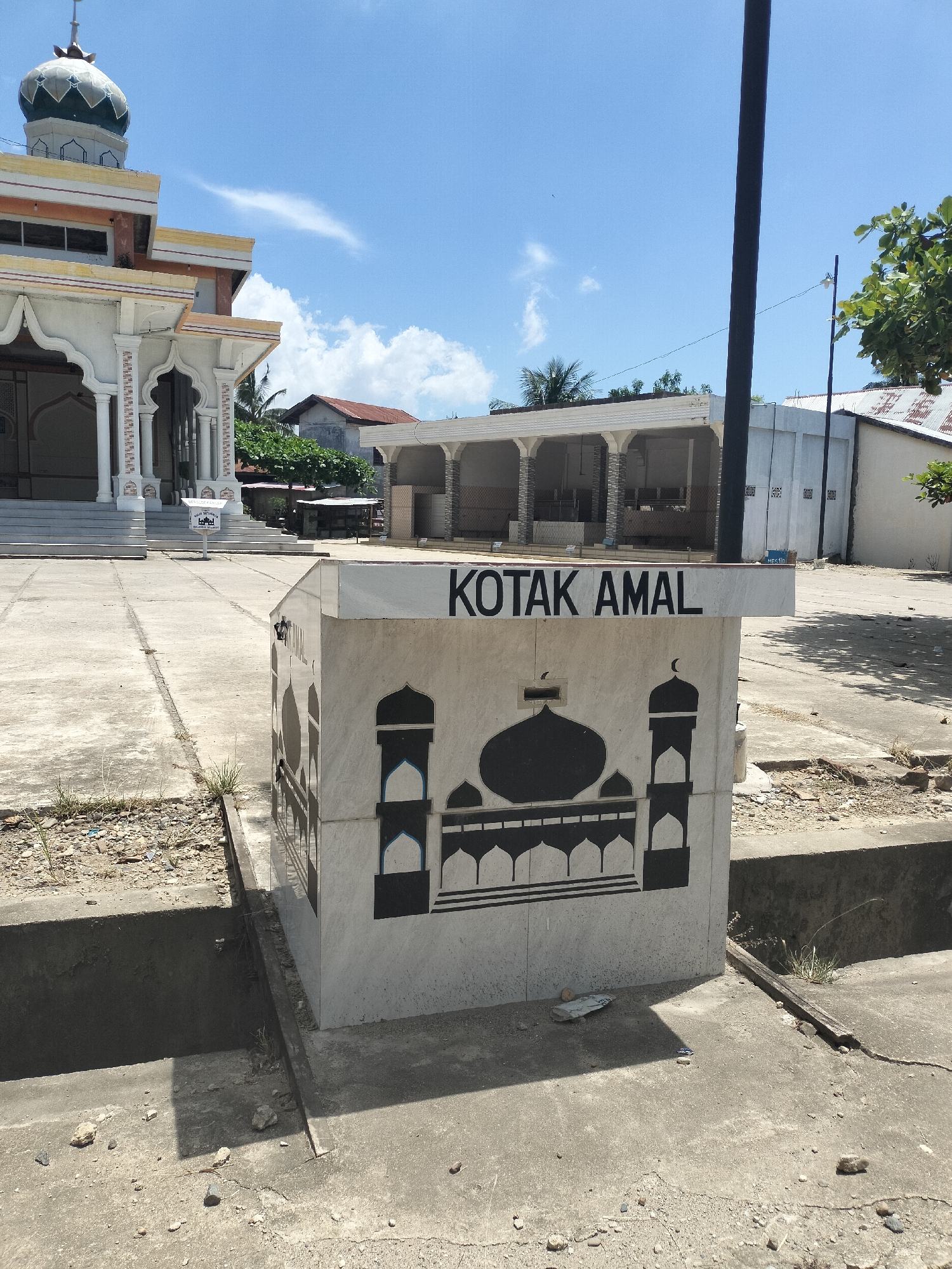 The image shows a donation box ("KOTAK AMAL") with a mosque illustration in front of a mosque in Matangkruet, Indonesia. The donation box is made of tiles and has a white background with black mosque imagery. The mosque in the background has a green dome and white walls with archways. The ground is paved with concrete. A black pole is present near the donation box. The sky is blue with a few clouds.