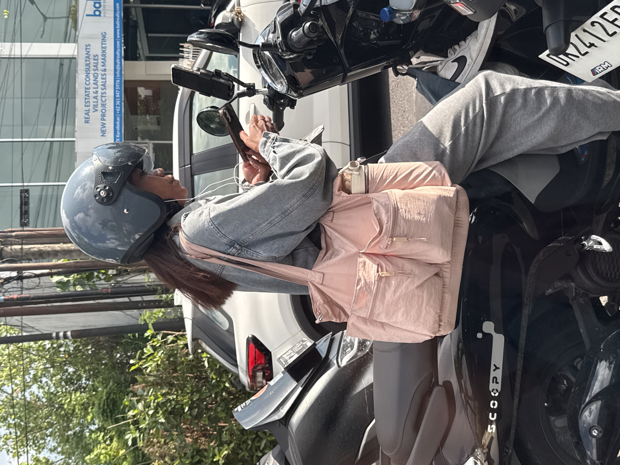 The image, oriented vertically, depicts a person seated sideways on a black Honda Scoopy scooter, stopped on a sunny street in North Kuta, Indonesia. The individual, appearing to be a woman with long dark hair, wears a grey full-face helmet, a light blue denim jacket, and grey sweatpants, paired with white sneakers featuring a black Nike swoosh.
She is actively engaged with her smartphone, holding it in both hands, and appears to be wearing wired earbuds. A pink shoulder bag is draped across her lap and the scooter's seat. Her posture is relaxed, with her left leg casually extended forward. A water bottle and a phone mount are visible on the scooter's handlebars.
In the background, a modern building with large windows stands tall. A prominent white sign on the building advertises "REAL ESTATE CONSULTANTS VILLA & LAND SALES NEW PROJECTS SALES & MARKETING" for "balirealty.com." The sign also includes contact information: "X Kerobokan | +62 361 847 5976 | info@balirealty.com | www.balirealty.com," explicitly pinpointing the location to Kerobokan, a district in North Kuta, Bali. Lush green trees and foliage are visible in the lower part of the background, contributing to a tropical setting. A white car is partially seen behind the scooter.
Further reinforcing the location, the scooter's license plate displays "DK 2412 FC," where "DK" is the regional code for Bali. The brand "SCOOPY" is clearly visible in white lettering on the side of the scooter. The bright sunlight and distinct shadows suggest it is daytime, likely mid-morning or afternoon.