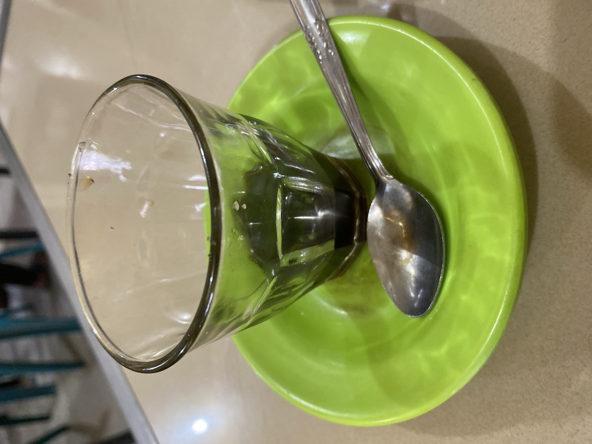 A close-up, overhead shot shows a clear glass with dark liquid, likely coffee, resting on a bright green saucer. A silver spoon with a patterned handle lies on the saucer, partially covering the glass. The setting appears to be a casual establishment, possibly a cafe or restaurant, with a table surface that is light brown and textured. In the blurred background, glimpses of teal chairs and a person's legs in dark pants suggest a public indoor space. The lighting is somewhat dim, with highlights reflecting off the glass and saucer. There is no indication of the specific location in Lhokseumawe, Indonesia, beyond it being an indoor dining setting.