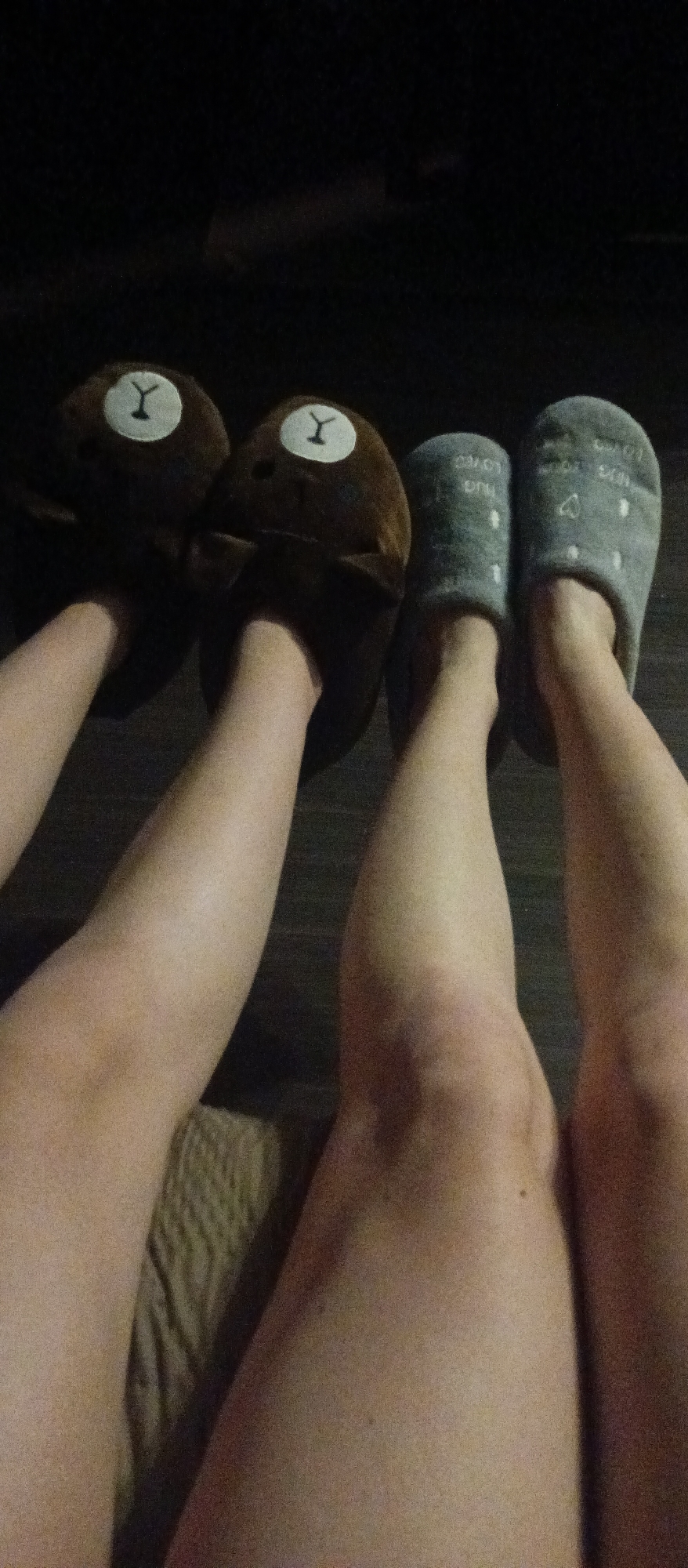 Two pairs of legs and feet are visible in a dimly lit indoor setting. On the left, a pair of legs stretches out, ending in feet clad in brown, bear-shaped slippers with white patches bearing a "Y" symbol. To the right, another pair of legs terminates in feet wearing grey slippers decorated with white dots and the words "hug" and "love." The legs are resting on what appears to be a patterned surface, possibly a couch or bedding. The background is dark and indistinct. The location of this scene within the city of Unknown, Unknown is not ascertainable from the provided image.
