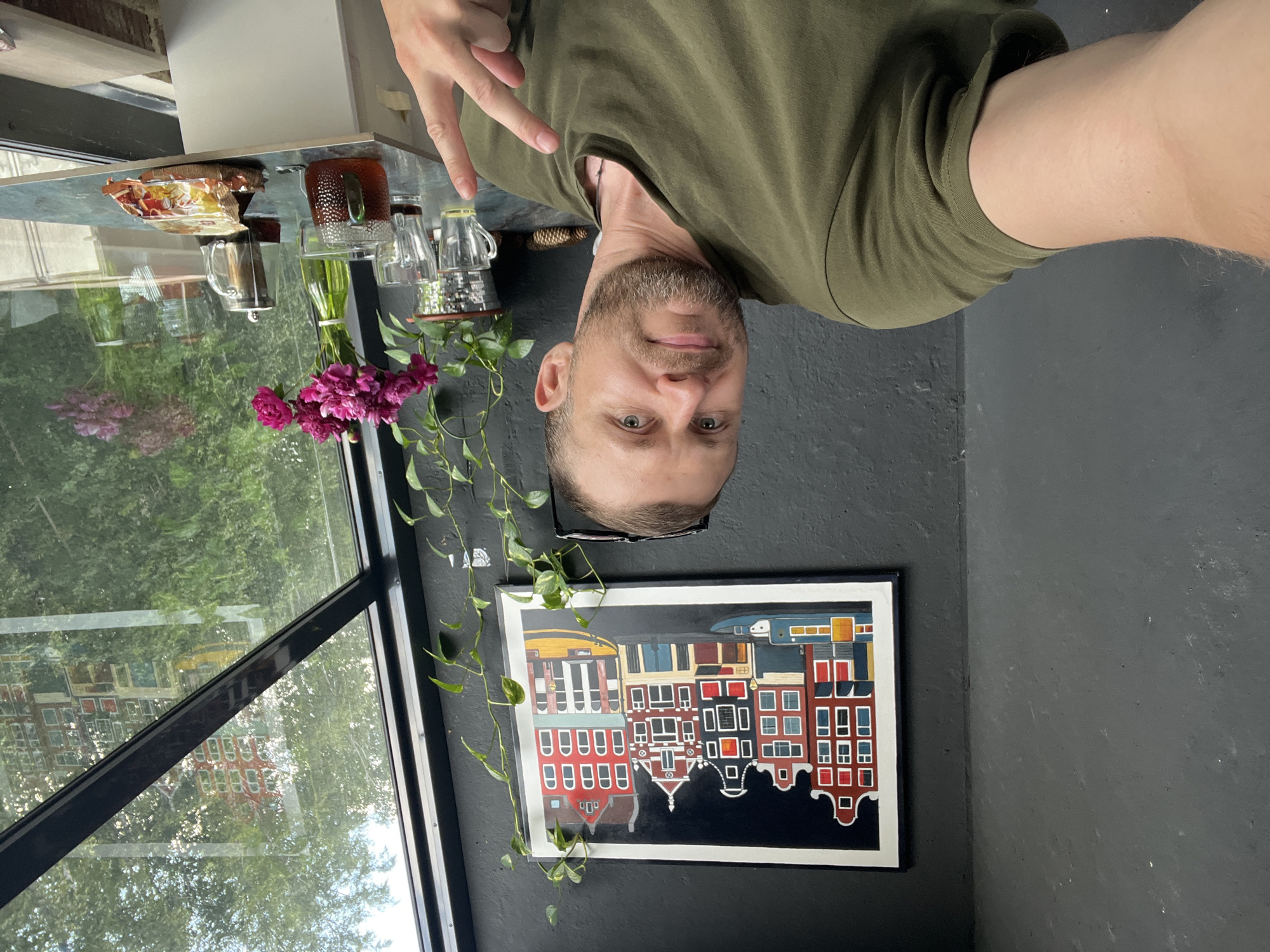 The image shows a man taking a selfie in an indoor setting. The man, with short brown hair and a beard, is wearing a green t-shirt and making a peace sign with his right hand. He appears to be smiling slightly.
On a glass-topped table in front of him, there are several objects: a package of what looks like biscuits, a glass mug with a brown tint, a small metal pitcher, two clear glass cups, and a green vase with pink flowers. A green vine with leaves drapes from the ceiling or a nearby shelf. Through the large window behind the table, lush green trees and foliage are visible, suggesting a pleasant outdoor environment, possibly a sunny day.
On the dark gray wall behind the man, a framed painting depicts a row of colorful, narrow buildings, characteristic of Dutch architecture. The overall scene suggests a relaxed moment, perhaps during a coffee break or a visit to a cafe. The lighting indicates it is daytime. There is no visible text in the image.