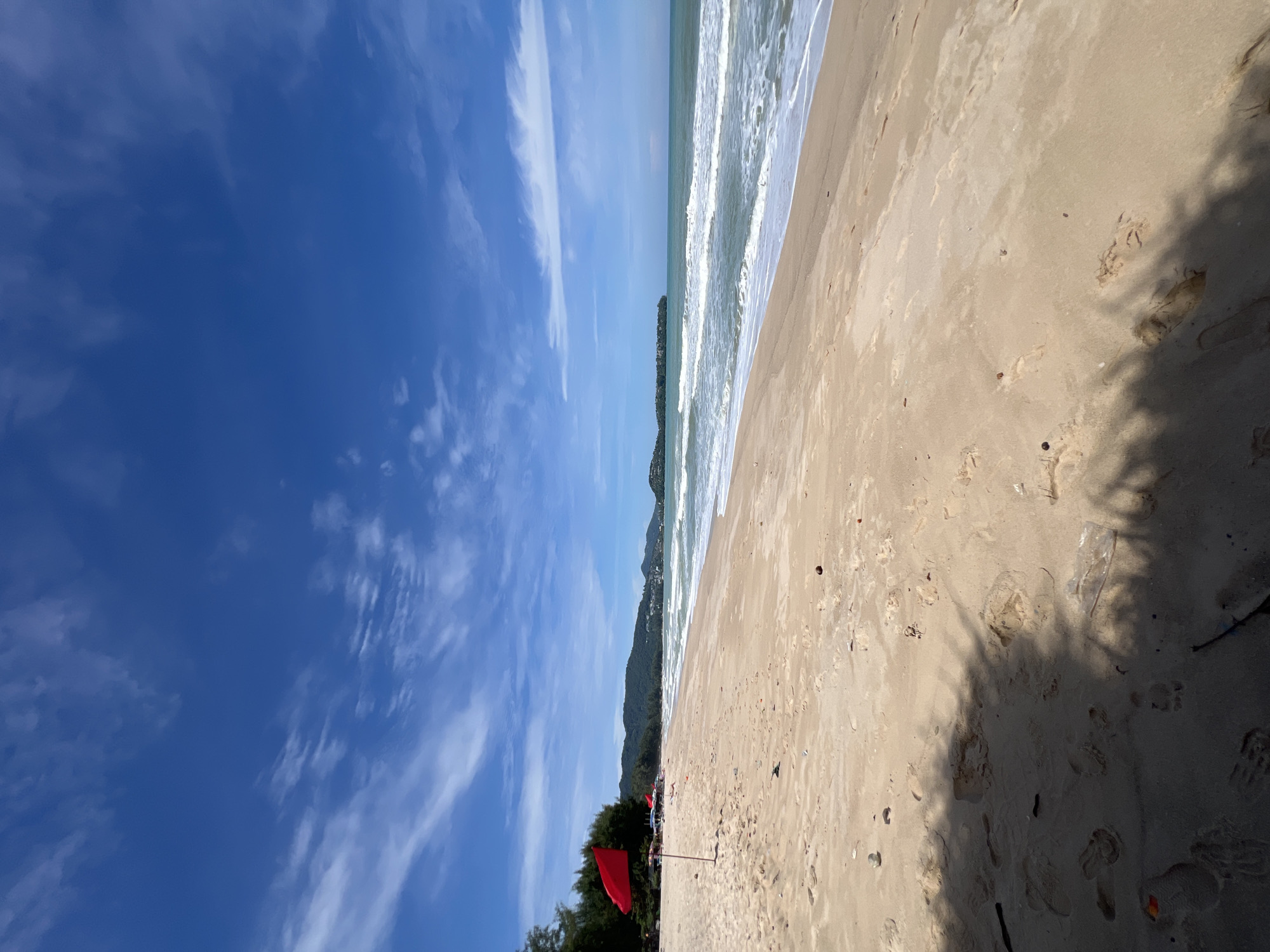 An eye-level, wide shot captures a tranquil beach scene under a bright blue sky dotted with wispy clouds. The sandy shore in the foreground is marked with footprints and scattered debris. A red flag stands prominently on the beach, likely a warning. Beyond the sand, gentle waves roll onto the shore from a calm, turquoise ocean. In the distance, a tree-covered coastline stretches out, with hills and buildings visible further down the shore. The overall impression is one of peacefulness and natural beauty.