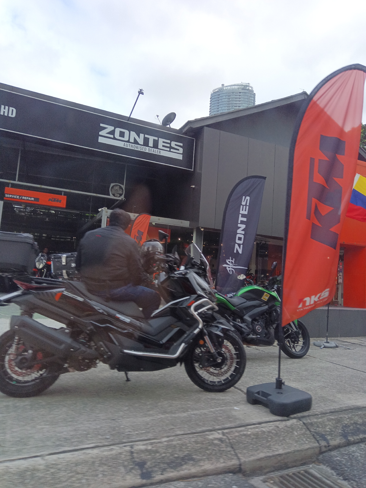 The image shows a street scene in Kuala Lumpur, Malaysia, in front of a motorcycle dealership. The dealership's signage indicates it sells ZONTES and KTM motorcycles. A large red flag with the KTM logo is prominently displayed. Several motorcycles are parked outside the dealership, including a black scooter in the foreground and a green motorcycle behind it. A person is sitting on the black scooter, facing away from the camera. The dealership appears to be located on a busy street, with a building in the background that resembles a high-rise apartment complex. The sky is overcast, suggesting a cloudy day.
