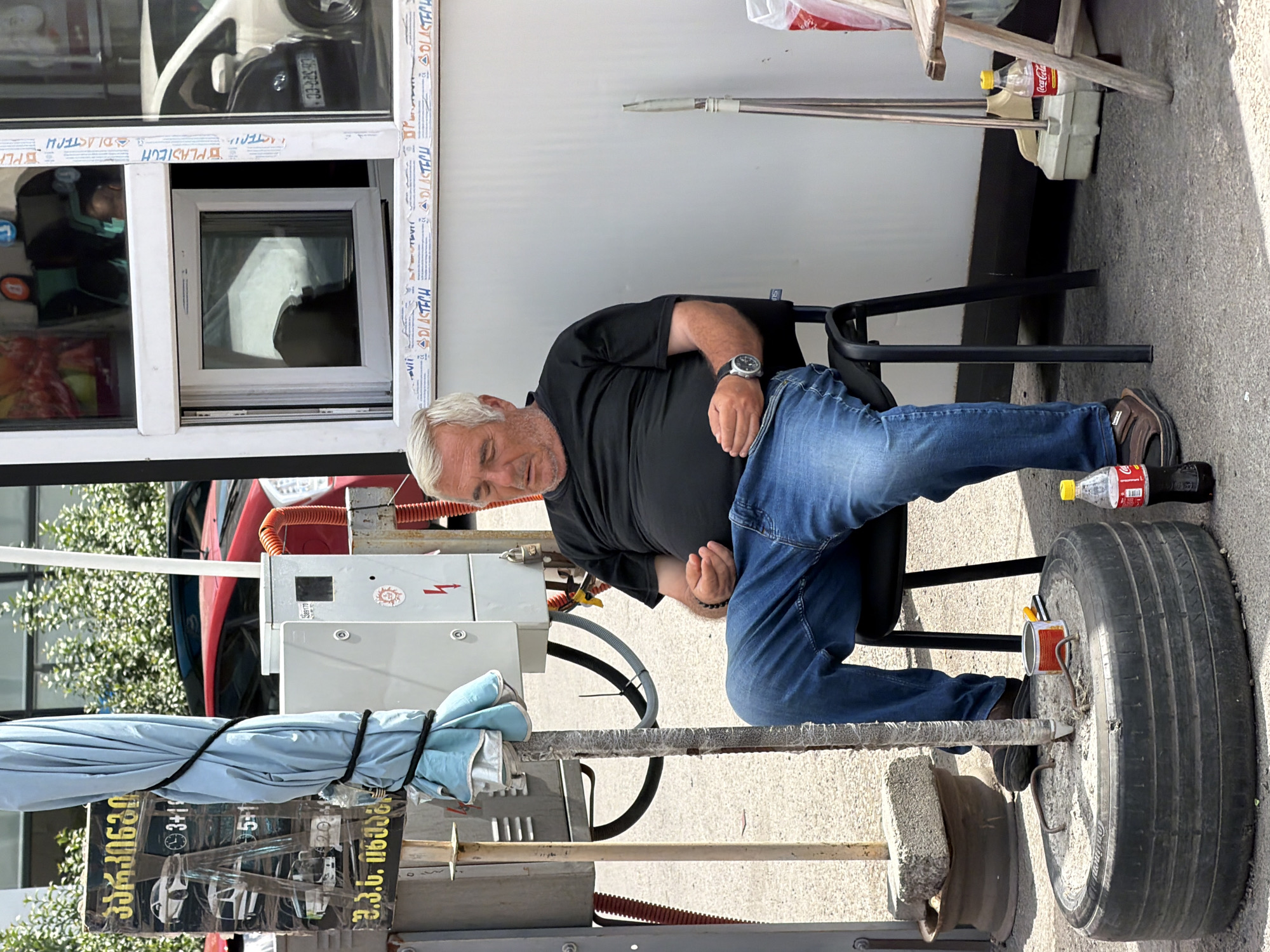 A middle-aged man with graying hair is sitting in a black plastic chair, his eyes closed, appearing to be asleep or resting. He is wearing a black t-shirt and blue jeans. A wristwatch is visible on his left wrist. The man is seated next to a large, worn tire that is filled with sand and used as a base for a metal pole, which is covered with a light blue tarp. A red and white can of food and a yellow-handled tool are resting on the sand within the tire. Next to the tire is a bottle of Coca-Cola.
Behind the man is a white structure with a window. The window is open, and a smaller window inside is partially obscured. The structure appears to be a kiosk or service booth. To the left of the man, a sign with Georgian writing and images of vehicles is attached to a metal pole. A metal box with electrical components and a padlock is mounted on a structure behind the man and to his left. The ground is paved with asphalt.
In the background, a red car is visible, and beyond that, green foliage. The scene suggests an outdoor, possibly semi-urban or commercial area, consistent with a location in Tbilisi, Georgia. The overall impression is one of a quiet, possibly hot day where the man is taking a break.