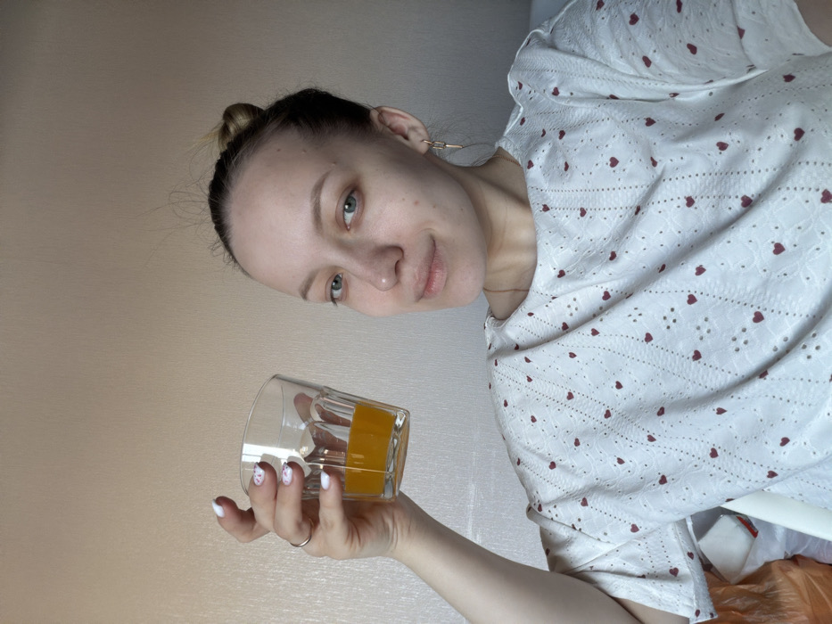 An indoor photographic capture in the city of Shipulino, Russia, depicts a female individual from the chest up. She possesses light skin, light-colored eyes, and her hair is styled in a bun with some loose strands. Her right hand is raised, holding a clear, faceted glass containing an orange liquid, approximately one-quarter full. Her fingernails are painted white with small red heart decals, and a silver-toned ring is present on her right ring finger. She wears a white short-sleeved top with an eyelet design featuring small red hearts and a thin gold-colored chain necklace. A gold-colored, rectangular earring is visible on her left ear. Her facial expression is neutral with a slight upward curve to the mouth, and she directs her gaze toward the viewer. The background is a plain, light-beige colored wall, with faint vertical texture visible. In the lower-left corner of the frame, portions of an orange plastic bag and a white object are discernible.