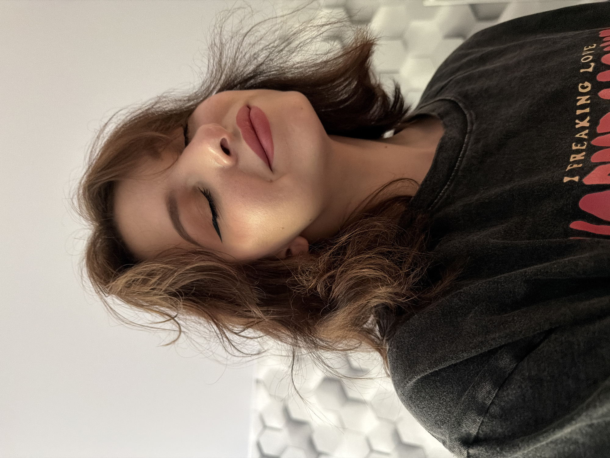 A young woman with medium-length brown hair, parted to the side, is shown in a close-up portrait. She is wearing a dark gray t-shirt with a graphic design on the front. Her eyes are closed, and her head is tilted slightly upwards. She has on subtle eye makeup with winged eyeliner and pink lipstick. The background is a wall with a textured, geometric pattern in shades of white and gray. The lighting suggests an indoor setting. There are no indications of the location being in a city.