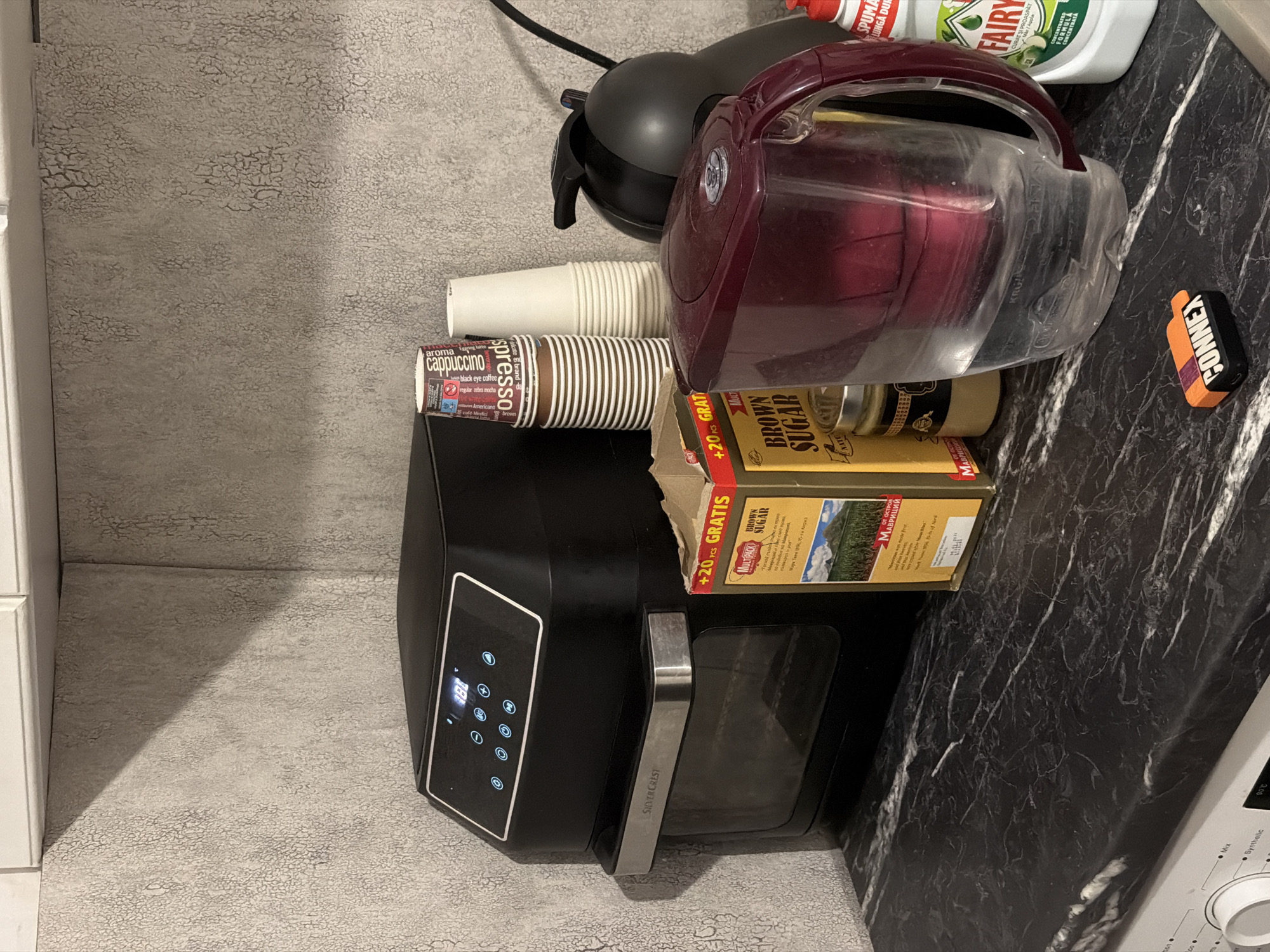 An indoor, eye-level, medium shot shows a kitchen counter with various appliances and food items. The wall behind the counter is textured and light gray, with a white cabinet visible at the top. The counter itself is black with white veining, resembling marble.
On the left, a black Silver Crest air fryer oven is positioned. Its digital display shows "181". To its right are stacked disposable coffee cups, some with "espresso" and "cappuccino" branding. Next to the cups is a box labeled "MultiPack BROWN SUGAR" with an image of a mountain landscape. A small glass jar with a yellow lid is placed next to the sugar box.
In the foreground, a large, clear plastic water filter pitcher with a dark red lid and handle is prominent. To the right of the pitcher, a bottle of "FAIRY" dish soap is visible. On the counter in front of the pitcher, a small, orange and black rectangular object with the word "CONNEX" is present.
The scene appears to be a domestic kitchen setting. Based on the presence of the "CONNEX" brand and its typical market presence, the location is likely in Plovdiv, Bulgaria.