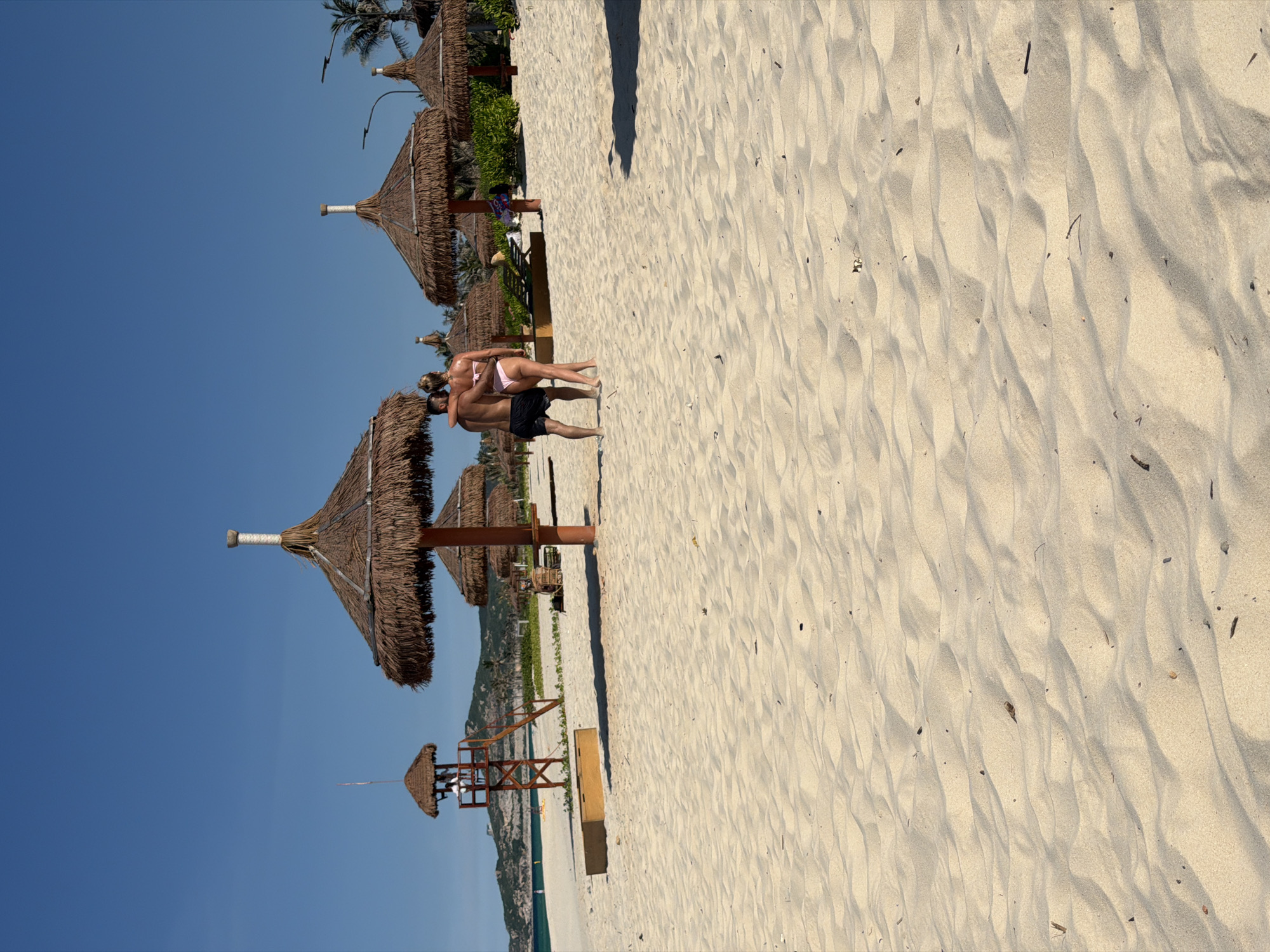 On a wide, sandy beach in Bac Cam Ranh Ward, Vietnam, a couple stands embracing near a cluster of thatched umbrellas. The man, wearing black swim shorts, has his arm around the woman, who is in a pink bikini. Behind them, several straw-covered beach umbrellas stand in a line, with some beach chairs visible further back. A lifeguard tower with a person in it is positioned to the left, and the turquoise ocean stretches out towards a distant, green-covered hill under a clear blue sky. The foreground of the image is dominated by the texture of the sand, showing ripples and a few scattered pieces of debris.