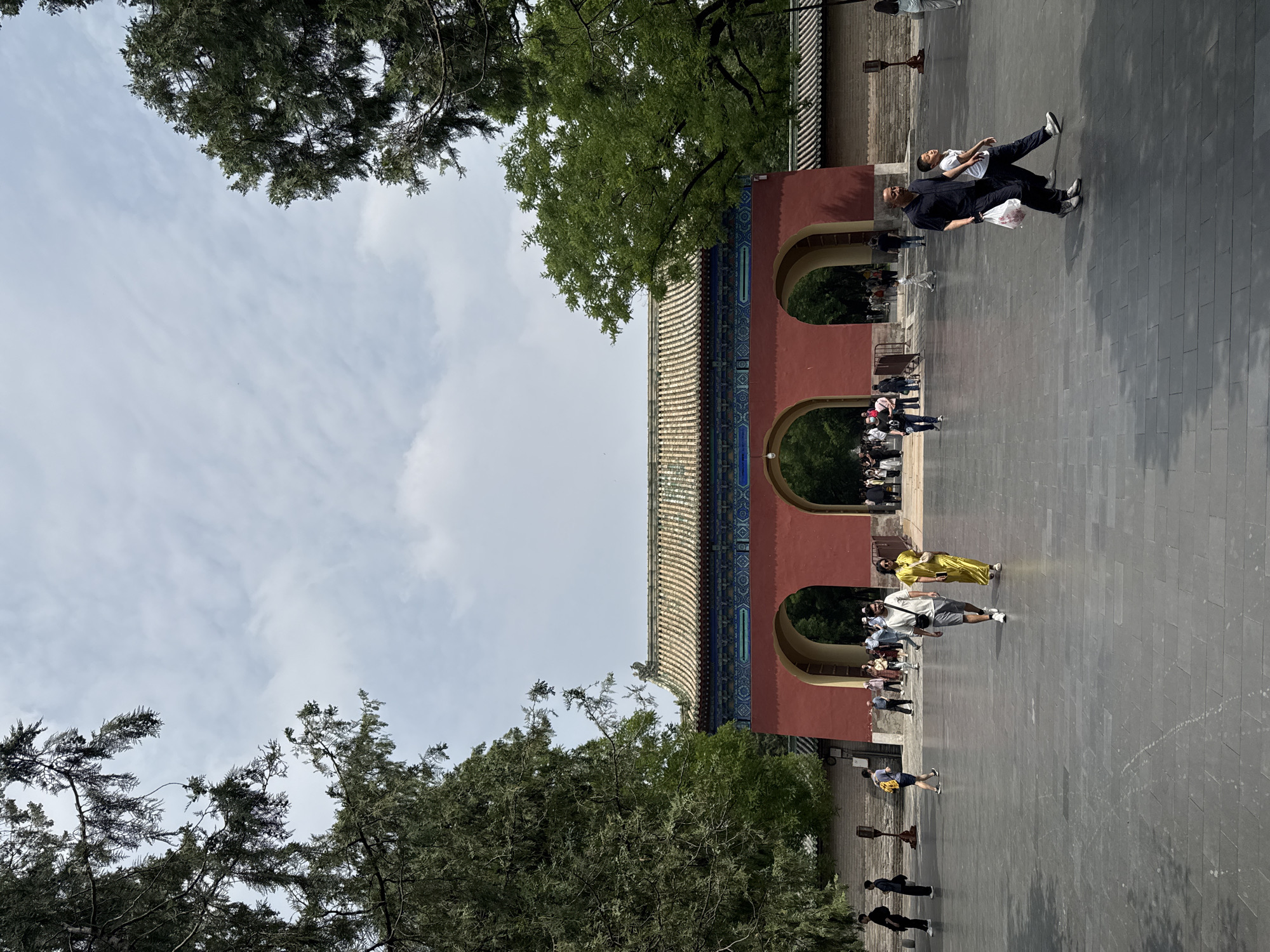 The image shows a red and tan traditional Chinese gate with three arched doorways, likely located in the Dongcheng District of China. The gate features a tiled roof and is surrounded by trees. People are seen walking in front of the gate, some of whom are wearing bright clothing. The environment is a paved area with trees visible in the background, under a cloudy sky.