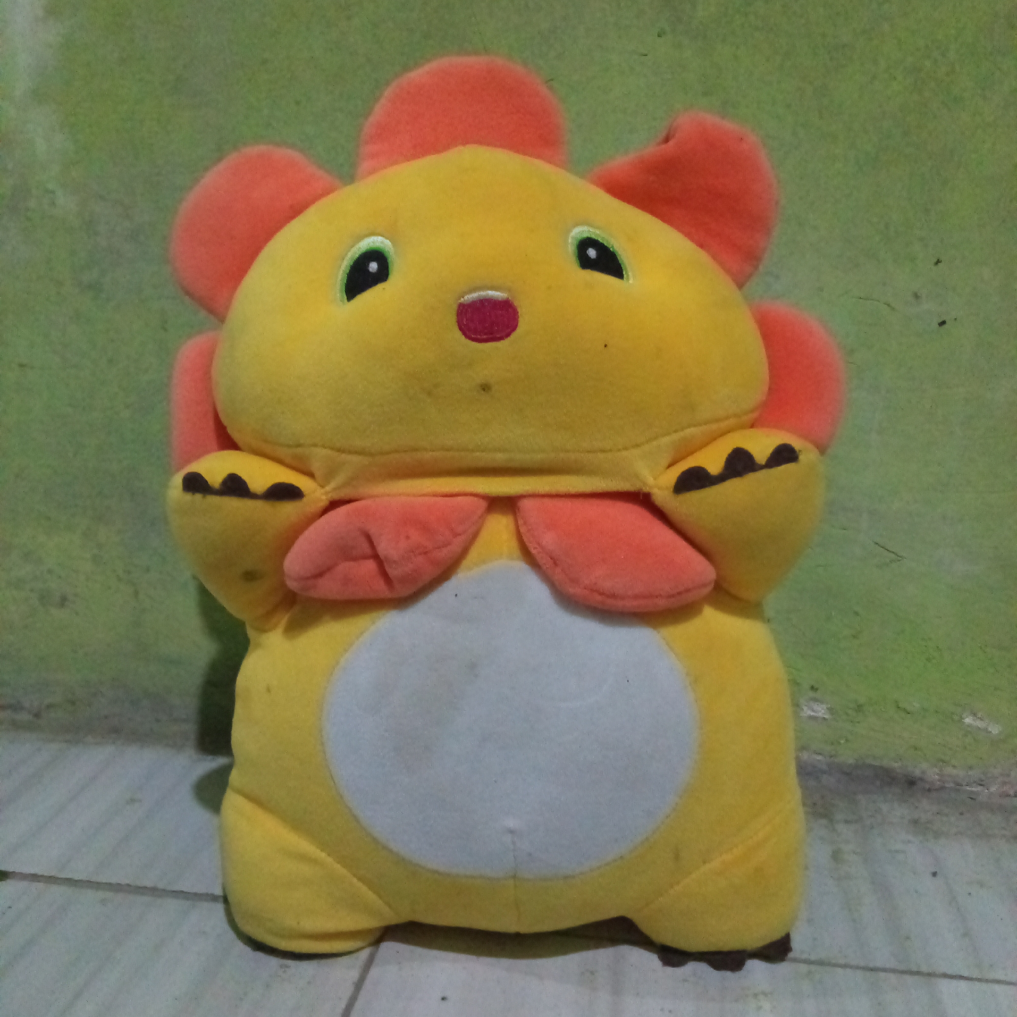 A close-up, slightly blurry, frontal view shows a yellow plush toy with orange accents. The toy is designed to resemble a character with a round head, large eyes, a small red mouth, and two large orange ears or petals on top. It has two small orange arms with black paw prints and two orange feet. A large, light-colored oval shape is present on the toy's belly. The toy is positioned on a light-colored tiled floor against a wall with a textured, faded green color. The overall scene appears to be an indoor setting, likely a room within a residence in Boyolali, Indonesia. There are no people visible in the image.