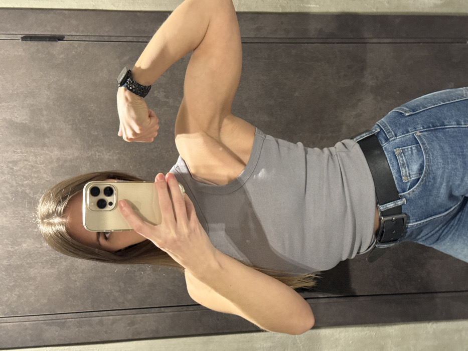 A woman poses for a mirror selfie. She is dressed in a gray tank top and blue jeans with a black belt. She flexes her bicep on her left arm, which also has a black wristwatch. She is holding a gold-colored iPhone. The background appears to be a wall or surface with a neutral gray tone.