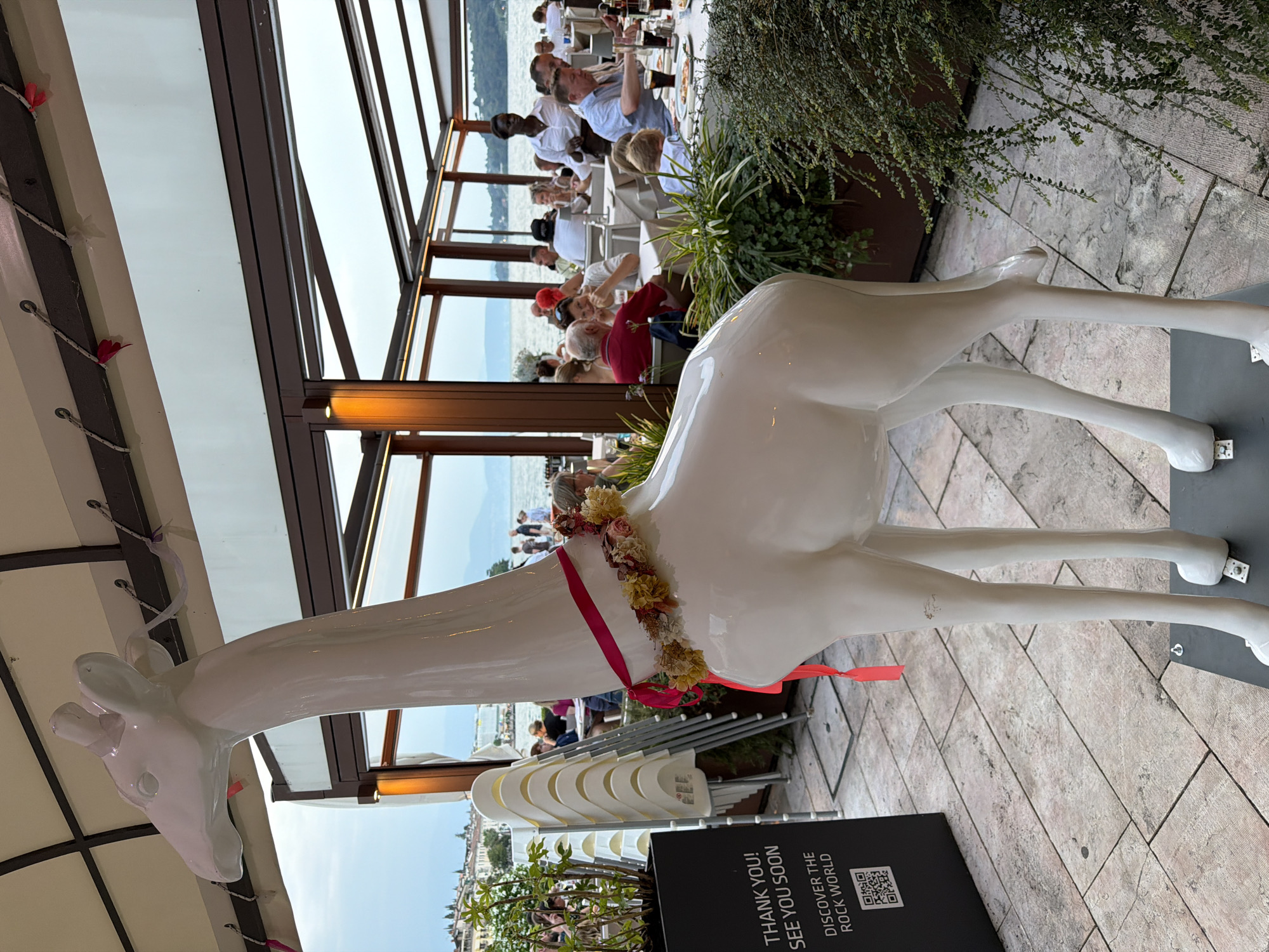 A white, life-sized giraffe statue stands on a tiled patio in front of an outdoor dining area of a restaurant situated in Salò, Italy. The statue is adorned with a pink ribbon and a floral garland around its neck. Behind the giraffe, patrons are seated at tables, some of whom are eating or conversing. A server, an African woman in a white shirt, is attending to the diners. The restaurant overlooks a body of water, likely Lake Garda, with hills and buildings visible in the distance under an overcast sky. A black planter with a QR code and text reading "THANK YOU! SEE YOU SOON. DISCOVER THE ROCK WORLD" is positioned to the left of the giraffe. Stacked chairs are visible near the planter, and lush greenery surrounds the patio.