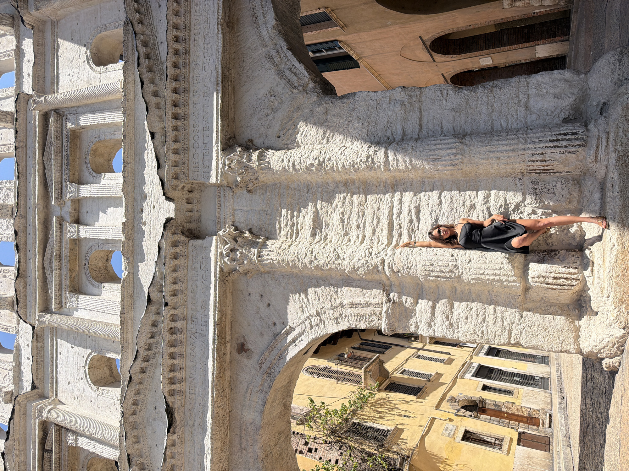 A low-angle shot captures a woman standing near a large stone archway in Verona, Italy, with her right hand raised against the textured stone pillar. She is wearing a black slip dress and sunglasses, and her left hand rests on her hip as she lifts the hem of her dress. The archway appears to be ancient, with weathered stone and visible inscriptions. To the left of the arch, a yellow building with shuttered windows and a balcony is visible, and a tree with green leaves grows against its facade. To the right, another building with arched doorways is seen. The scene is bathed in bright sunlight, casting strong shadows. The inscription on the archway translates to "Honor and Glory to Augustus, Son of the Divine Julius."