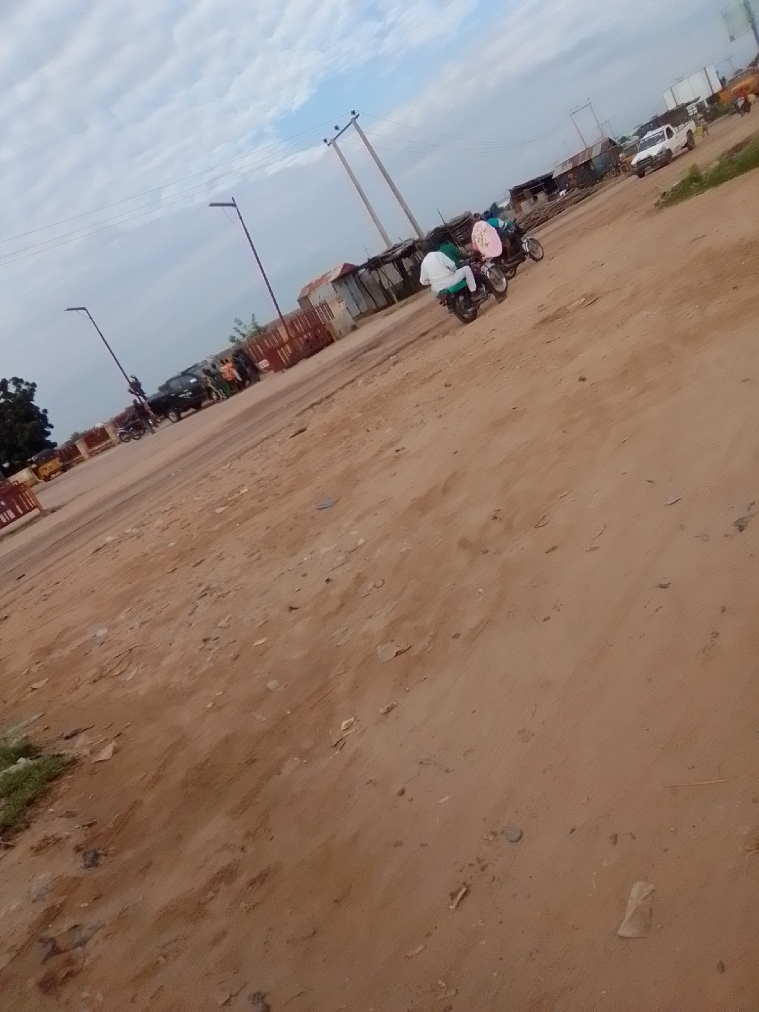 The image depicts a dusty, unpaved road in what appears to be a peri-urban or rural area, consistent with parts of Gombe, Nigeria. The scene is captured from a low angle, looking across the expanse of reddish-brown dirt towards a collection of buildings and a few vehicles. The sky is overcast with scattered clouds.
In the foreground, the dusty ground is uneven with some scattered debris. A person in a white garment is seen on a motorcycle in the middle ground, facing away from the camera. Another motorcycle, with a rider wearing a green garment and a passenger in a pink garment, is positioned slightly behind and to the left of the first. The motorcycles appear to be moving or stopped on the dirt road.
Further back, several small, makeshift structures with corrugated metal roofs are visible, suggesting a commercial or residential area under development or informal settlement. A white pickup truck is parked on the right side of the road, and other vehicles, including what looks like a yellow tricycle (Keke Napep), are present in the distance, though partially obscured. A few individuals are gathered near the structures. Utility poles with wires are visible in the background, indicating the presence of infrastructure. The overall impression is of a busy, active, but somewhat underdeveloped area in Gombe.