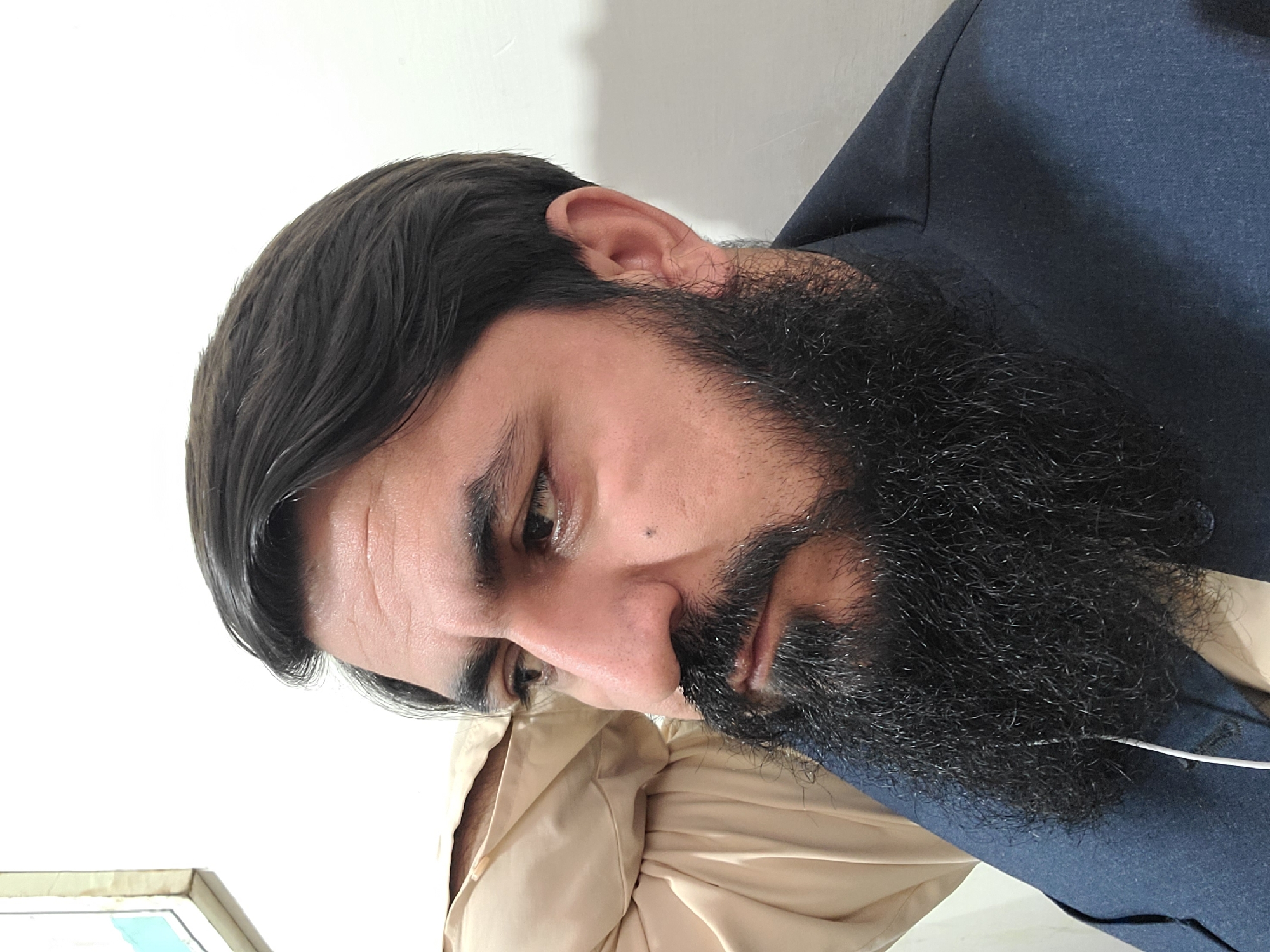 A headshot depicts a man with dark hair and a full black beard, possibly in Quetta City Tehsil, Pakistan. He has a slight frown. He is wearing a dark blue suit over a light tan shirt.