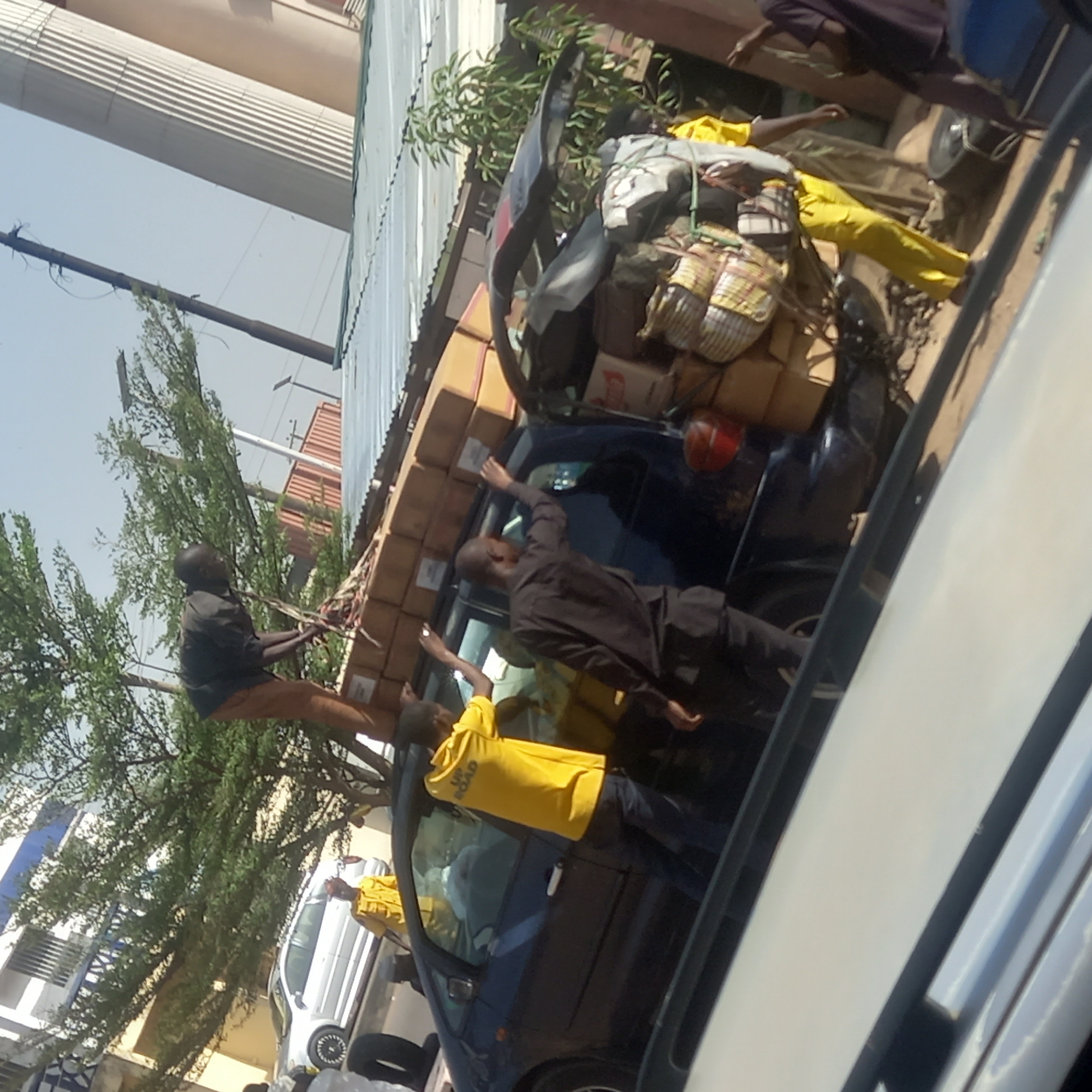 The image depicts an outdoor scene, likely on a street or in a market area in Sardauna, Nigeria, during bright daylight. Several men are engaged in the process of loading or securing a large quantity of goods onto the roof of a dark-colored vehicle, possibly a hatchback or small SUV.
At least four men are visible. One man, dressed in a dark shirt and light-colored pants, is perched on the side of the car near its front, appearing to arrange or tie down items. Another man, wearing a black long-sleeved shirt and dark pants, stands alongside the vehicle, reaching up towards the cargo. A third man, sporting a yellow t-shirt with "UP ROAD" printed on it and dark pants, is also standing next to the car, assisting with the loading. A fourth person, partially visible towards the rear of the vehicle, also wears a yellow outfit.
The vehicle is heavily laden with numerous cardboard boxes stacked high on its roof. Interspersed with the boxes are bundles wrapped in patterned, possibly plaid or checkered, fabric, suggesting luggage or other merchandise. These items are secured with ropes or straps. A red Coca-Cola box is identifiable among the cargo.
In the background, a large building with distinct light-colored, ribbed architectural features (possibly columns or corrugated siding) rises against a clear, bright blue sky. Lush green trees are visible, and a utility pole or structure stands in the distance. The presence of another white car partially visible further down the street suggests an urban or semi-urban setting. The scene highlights the common practice of transporting goods with vehicles heavily loaded, indicative of a bustling commercial or transit activity.