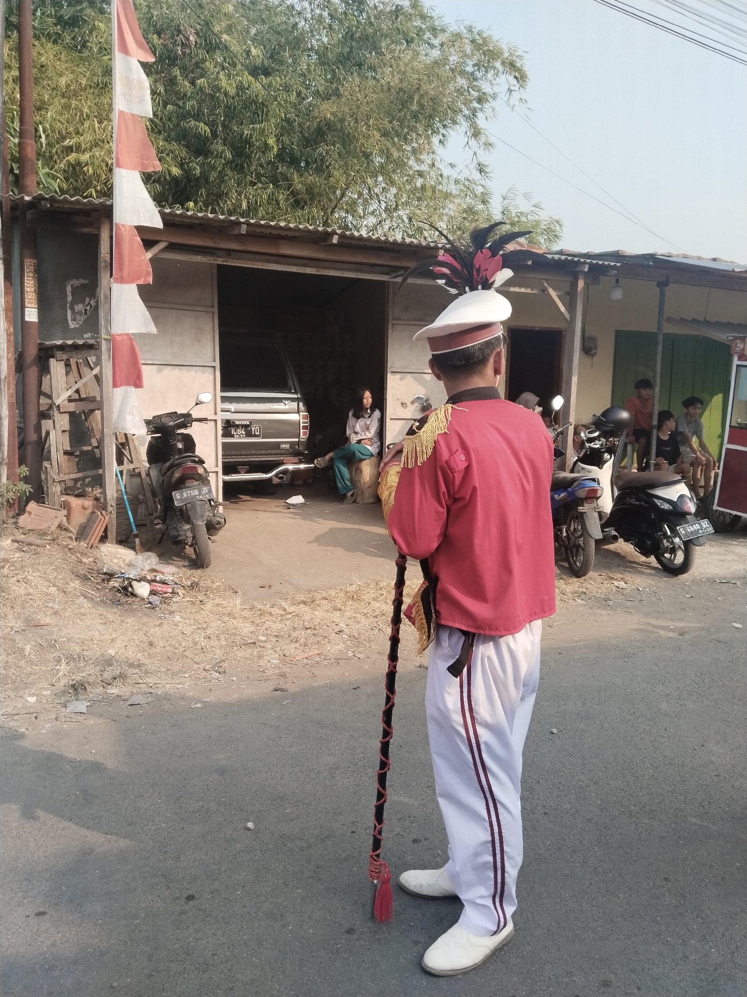 The image depicts a street scene in Margasari, Indonesia. In the foreground, a person wearing a red marching band uniform with a white hat adorned with feathers stands with their back to the camera, holding a baton. The uniform includes white pants with red stripes. Behind them, several motorcycles are parked. A building with a corrugated metal roof and an open front is visible, with a black car parked inside. A woman is seated on a wooden stool to the right of the building. Further back, several children are visible, some sitting on a bench and others standing. A flag with alternating red and white stripes hangs from a pole on the left side of the image. The ground is paved with asphalt, and the surrounding area appears to be a mix of dry vegetation and debris. The sky is clear and bright, suggesting daytime.