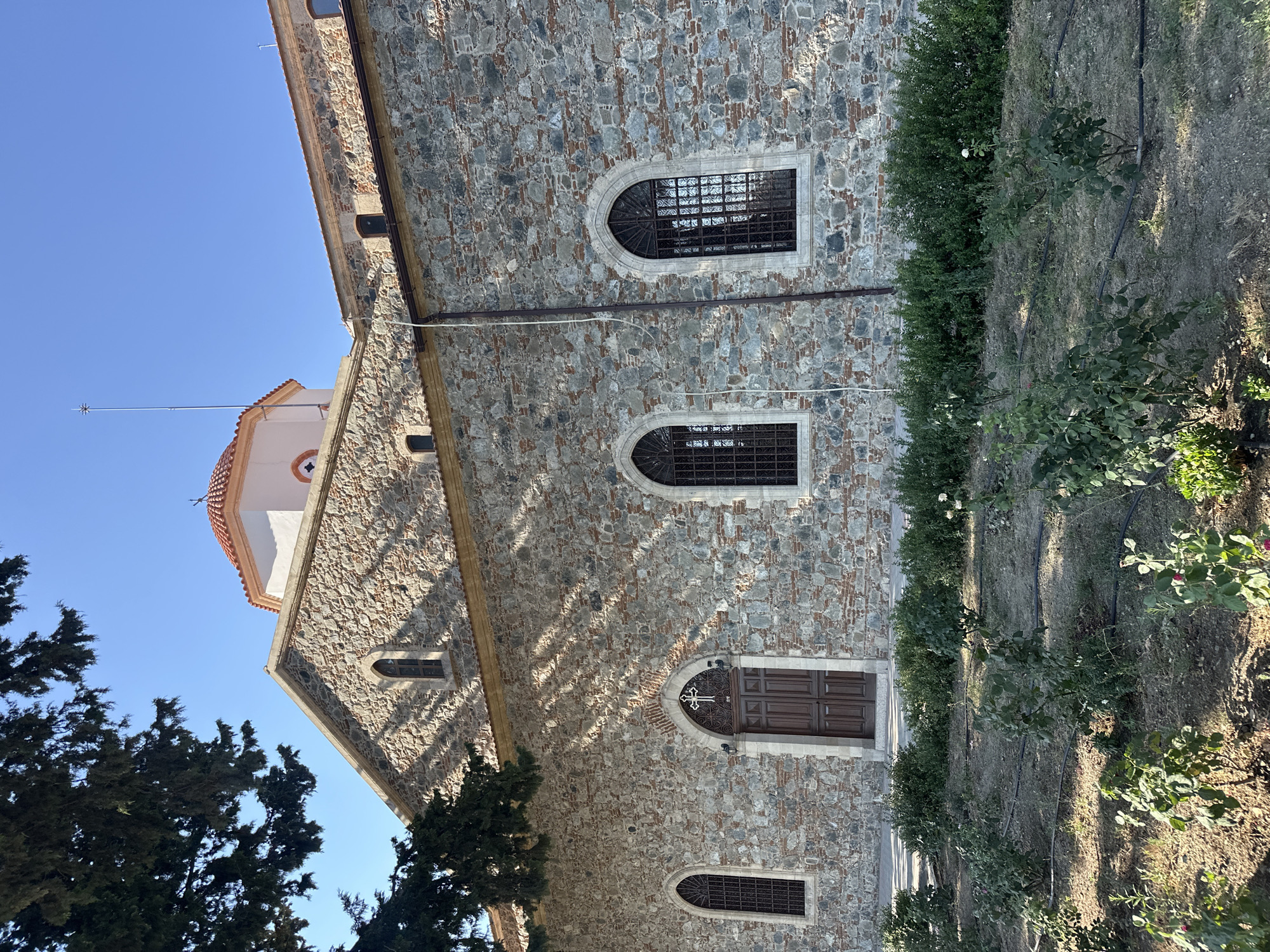 The image captures a close-up, eye-level, exterior view of a stone-built church in Agros, Cyprus, on a clear, sunny day. A large tree with dark green foliage extends into the upper left corner, casting dappled shadows across the building's facade. The church walls are constructed from rough-hewn stones and bricks, with arched windows featuring dark metal grilles. A prominent wooden double door with a cross motif above it is centrally located on the main facade. The roofline is partially visible, with a section of a light-colored structure topped by a red-tiled dome. In the foreground, a garden area with young rose bushes and visible irrigation lines occupies the lower portion of the frame. The sky is a bright, cloudless blue.