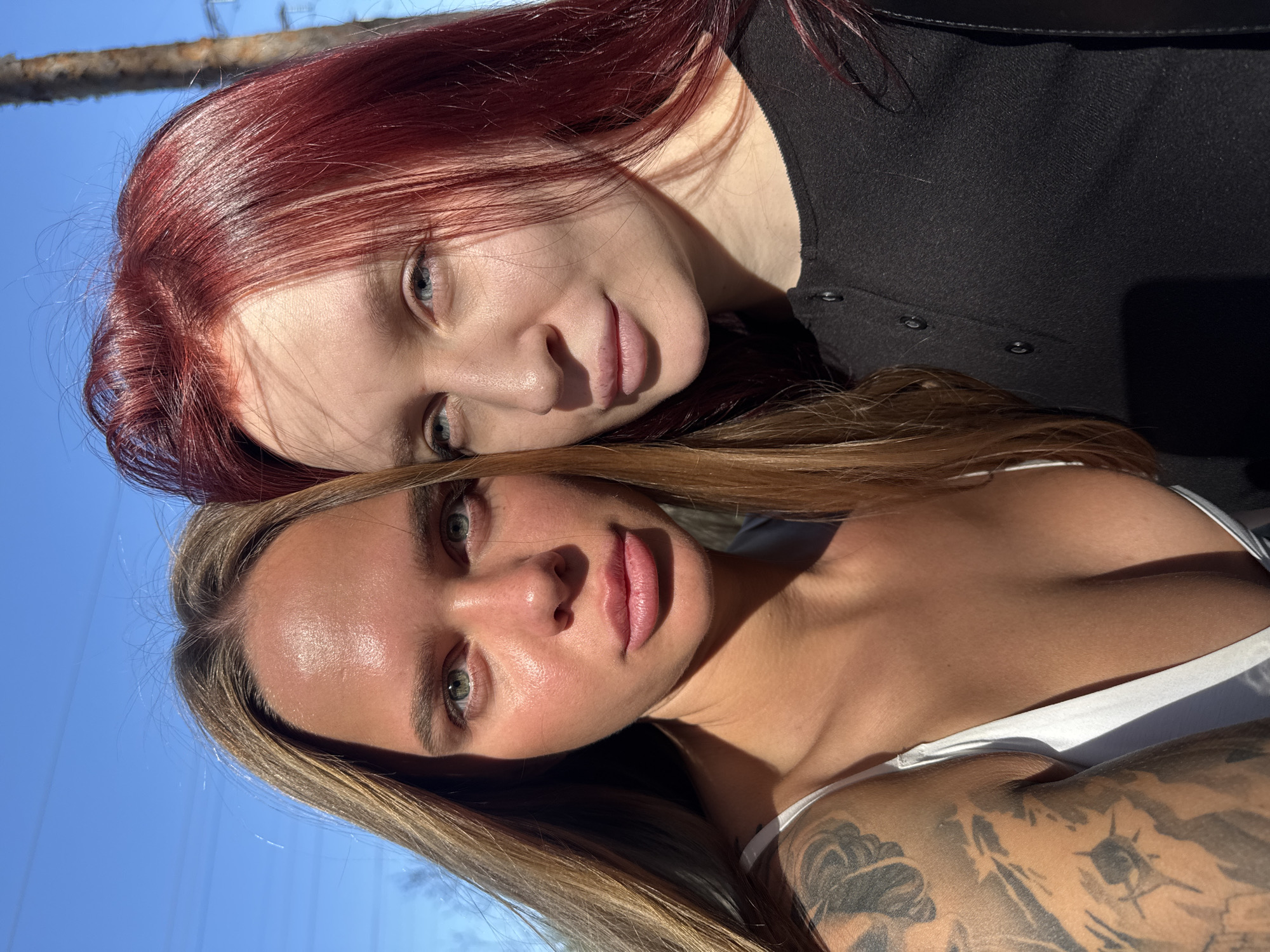 Two women are closely positioned, likely posing for a photograph. The woman in the foreground has blond hair, green eyes, and a light complexion. She is wearing a white top, and a tattoo is visible on her arm. The other woman, partially behind the first, has red hair, blue eyes, and is wearing a black top with buttons. The background shows a clear blue sky and a wooden post or tree branch at the top of the frame. The image suggests an outdoor setting with good lighting. The photo's location in the city of Nizhny Novgorod, Russia, cannot be confirmed without additional information.