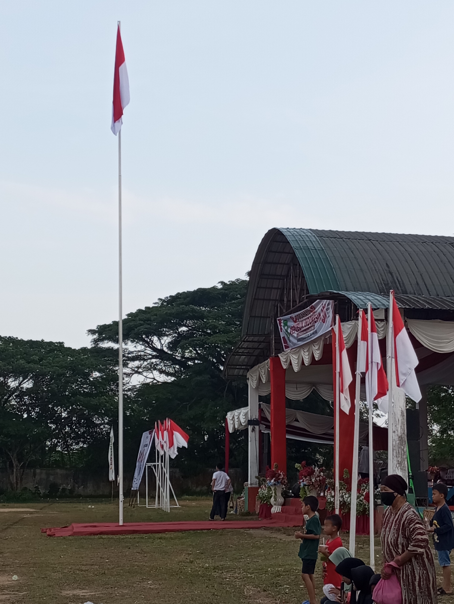 In Candirejo, Indonesia, a flag-raising ceremony is underway, marked by the Indonesian flag, a red and white banner, hoisted on a tall pole. The ceremony takes place in an open field adjacent to a covered stage. Several Indonesian flags are displayed on poles in front of the stage. The stage is decorated with red and white fabric and flowers. People, including children and adults, are gathered to witness the event. Some are standing, while others are seated on the ground. A woman in a patterned dress and headscarf, wearing a mask, is observing the ceremony. The environment includes trees in the background and a cloudy sky overhead.