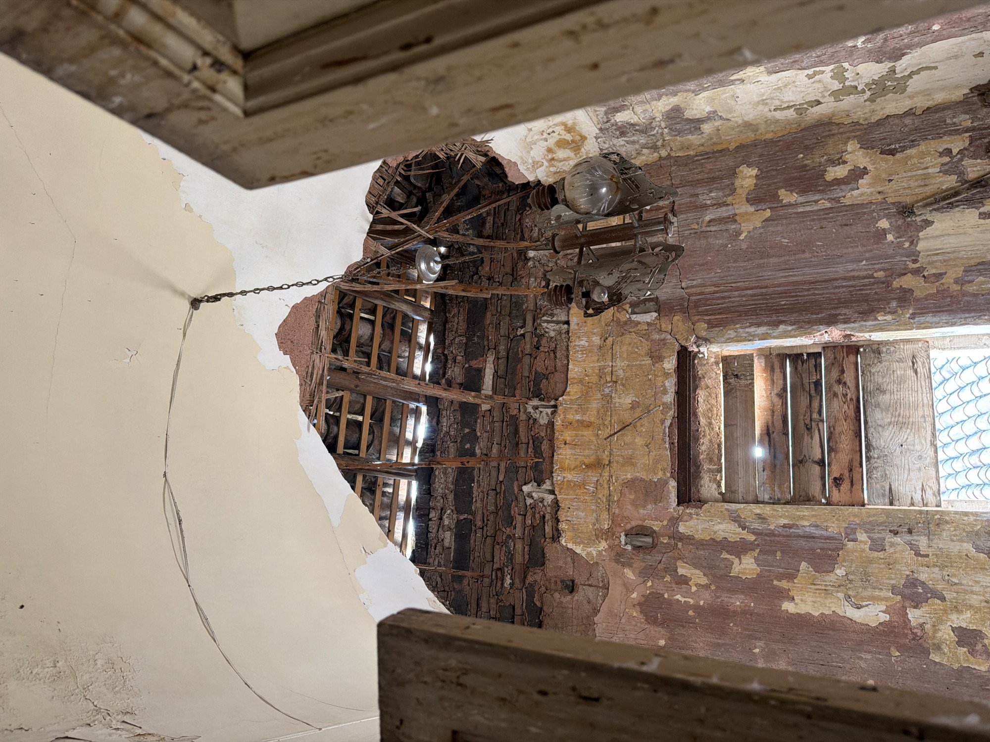 The image presents the interior of a severely dilapidated room within a building located in Catania, Italy. The perspective is a low-angle shot, looking upwards towards the ceiling and walls.
The arched ceiling, originally plastered, exhibits extensive damage, with large sections having collapsed. Exposed wooden roof beams and lathing are visible through the gaps, with bright sunlight penetrating from above, suggesting a partial roof collapse or missing roof tiles. The remaining plaster on the ceiling is light cream-colored, showing significant cracking, water damage, and peeling paint. A dark, thin cable runs along a crack in the ceiling.
Below the ceiling, the walls are in an advanced state of decay. Multiple layers of paint, primarily faded reddish-brown and yellow-ochre over lighter undercoats, are extensively peeling and flaking. Higher portions of the walls reveal rough, dark-colored exposed brickwork where plaster has completely deteriorated.
A prominent, aged metal chandelier, likely rusted, is suspended by a chain from a point of damage in the ceiling, slightly right of center. The chandelier features ornate, possibly scrollwork, elements and contains two visible clear glass light bulbs. To the left and slightly higher than the main chandelier, a simpler light fixture, appearing as a bare bulb or reflector, hangs by a wire.
Towards the bottom center of the frame, a rectangular opening, consistent with a window, is boarded up horizontally with five weathered wooden planks. Bright sunlight filters through the gaps between these planks. Below the wooden boards, the upper edge of a terracotta tiled roof is visible, indicating an exterior view over an adjacent structure. On the far left and right edges of the image, portions of aged, unpainted wooden architectural elements, possibly door jambs or structural supports, frame the scene. No individuals or active actions are present, conveying a scene of profound abandonment and structural neglect.