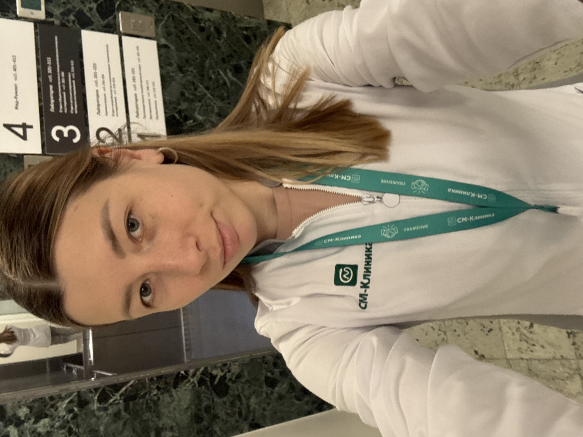 A young woman in a white, long-sleeved top or jacket is captured in a selfie. She has light brown hair and is looking directly at the camera with a neutral to slightly pleasant expression. She wears a hoop earring on her left ear. Around her neck is a teal-colored lanyard featuring the text "СМ-Клиника" (SM-Klinika), its logo (a stylized 'S' and 'M'), and the word "УВАЖЕНИЕ" (RESPECT). The same "СМ-Клиника" text and logo are also embroidered on the left chest area of her white top.
The setting appears to be an indoor environment, likely a medical clinic or office building in Moscow, Russia, given the Cyrillic text. The background features a dark green, marble-patterned wall. On the wall, several black and white signs are visible, indicating different floor levels and departments. These signs read "4 Мед-Регист каб. 401-413" (Med-Regist office 401-413), "3 Лаборатория каб. 300-313" (Laboratory office 300-313), and "2 Лабаратория каб. 200-223" (Laboratory office 200-223). A partially obscured sign for a "Лаборатория" (Laboratory) on floor "1" is also visible. The woman's reflection, also wearing a white top, can be faintly seen in a reflective surface behind her to the left, suggesting a mirror or elevator panel. The time of day is not discernible.