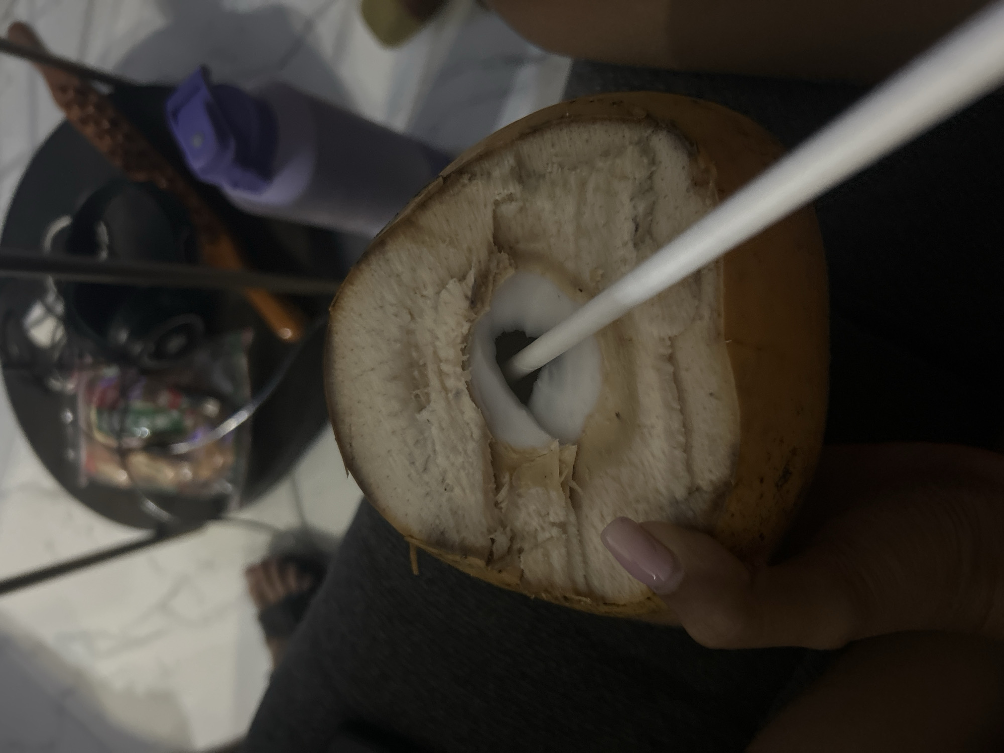 The media file features a close-up shot of a person holding a halved coconut with a white straw inserted into it. The coconut appears fresh, with its white flesh visible. The person's hand, with a light pink nail polish, is gently holding the coconut. In the background, out of focus, are various objects including what appears to be a dark-colored hairbrush with a purple cap, and other items on a shelf or table, possibly in a kitchen or indoor setting. The lighting suggests it is daytime. The scene is likely in Sukawati, Indonesia, as indicated by the location context. The overall impression is one of refreshment and tropical enjoyment.