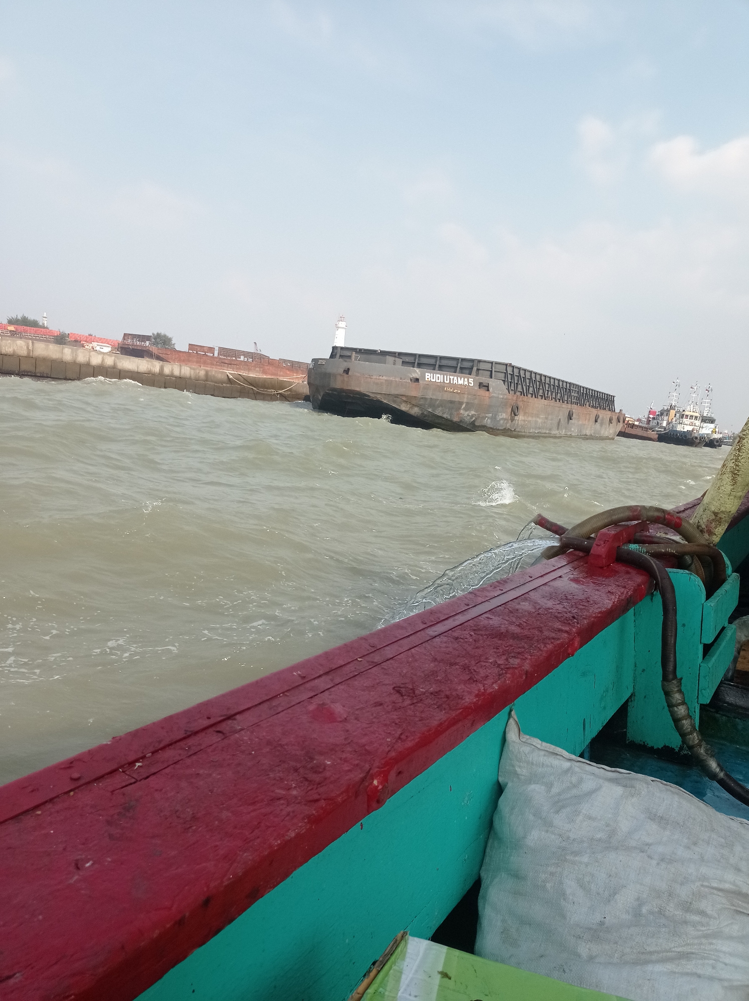 A high-angle, wide shot captures a body of water in Tegal, Indonesia, with several ships visible. In the foreground, the deck of a boat is visible, with a red railing and teal-colored sides. A white sack is leaning against the side of the boat. In the mid-ground, a large barge labeled "BUDI UTAMAS" is partially submerged in the water. Further in the distance, another barge is moored to a concrete pier with a lighthouse. Several tugboats are also visible near the pier. The water is choppy, and the sky is overcast.