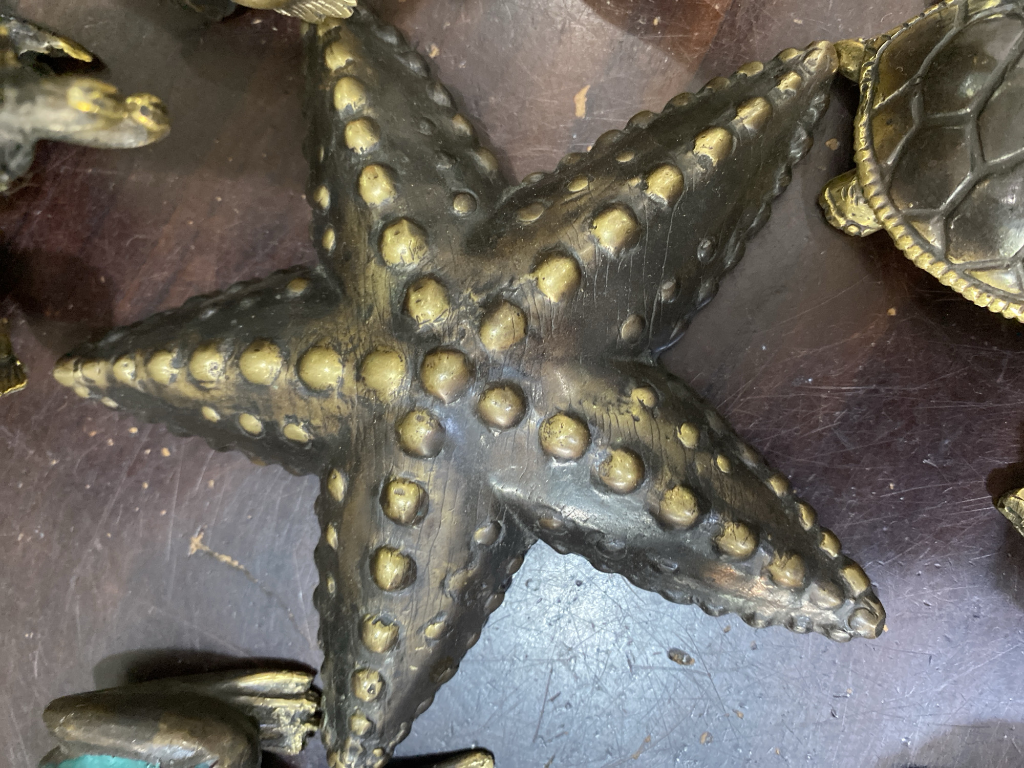 A high-angle, close-up shot depicts a dark metallic starfish, likely a decorative object, lying on a dark, possibly wooden, surface. The starfish has five arms and is textured with numerous raised, rounded bumps. Partial views of other dark metallic objects, including what appears to be a turtle figure, are visible near the edges of the frame. The environment suggests an indoor setting, possibly a shop or display area. The image does not contain any people. The location in Nusa Dua, Indonesia, is not evident from the image.
