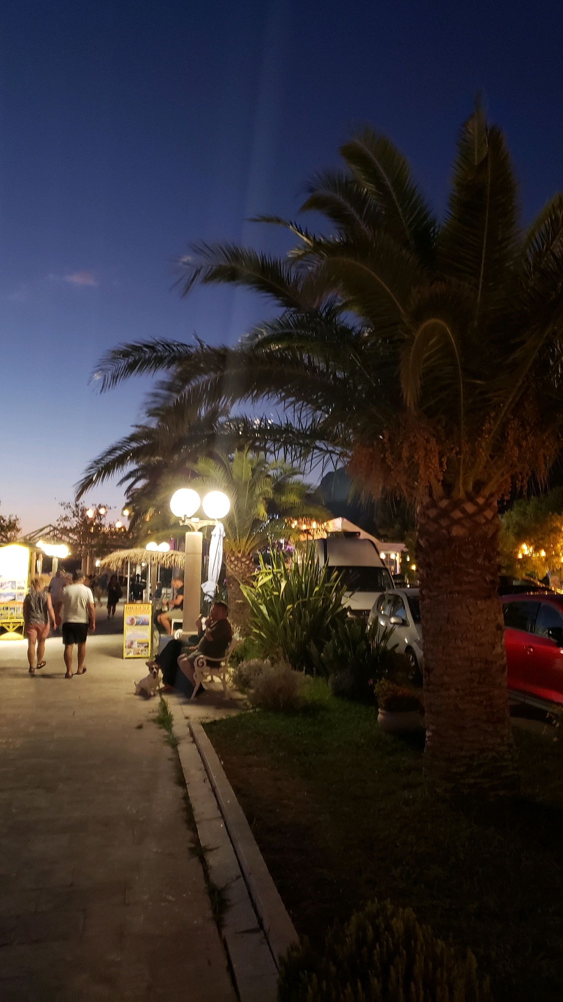 The image depicts a scene at dusk in Općina Tučepi, Croatia. A paved walkway lines the left side of the frame, with several people walking and standing. In the foreground, a couple walks away from the camera, with the woman in a patterned top and pink shorts, and the man in a white t-shirt and dark shorts. Further down the walkway, a person sits on a bench next to a small white dog, appearing to be smoking. Several palm trees are visible throughout the scene, some with illuminated fronds, suggesting the presence of outdoor lighting. Behind the walkway, there are market stalls or kiosks with lighting and signs. A white van and a silver car are parked further back, and a red car is visible on the far right. The sky is a deep blue, transitioning to a lighter hue near the horizon, indicating the evening hours. The overall atmosphere is relaxed, possibly a promenade area by the coast.